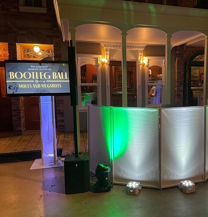 A DJ booth setup at a wedding or party, with colorful lighting, a screen inviting guests to a 'Bootleg Ball,' and a wooden house facade background.