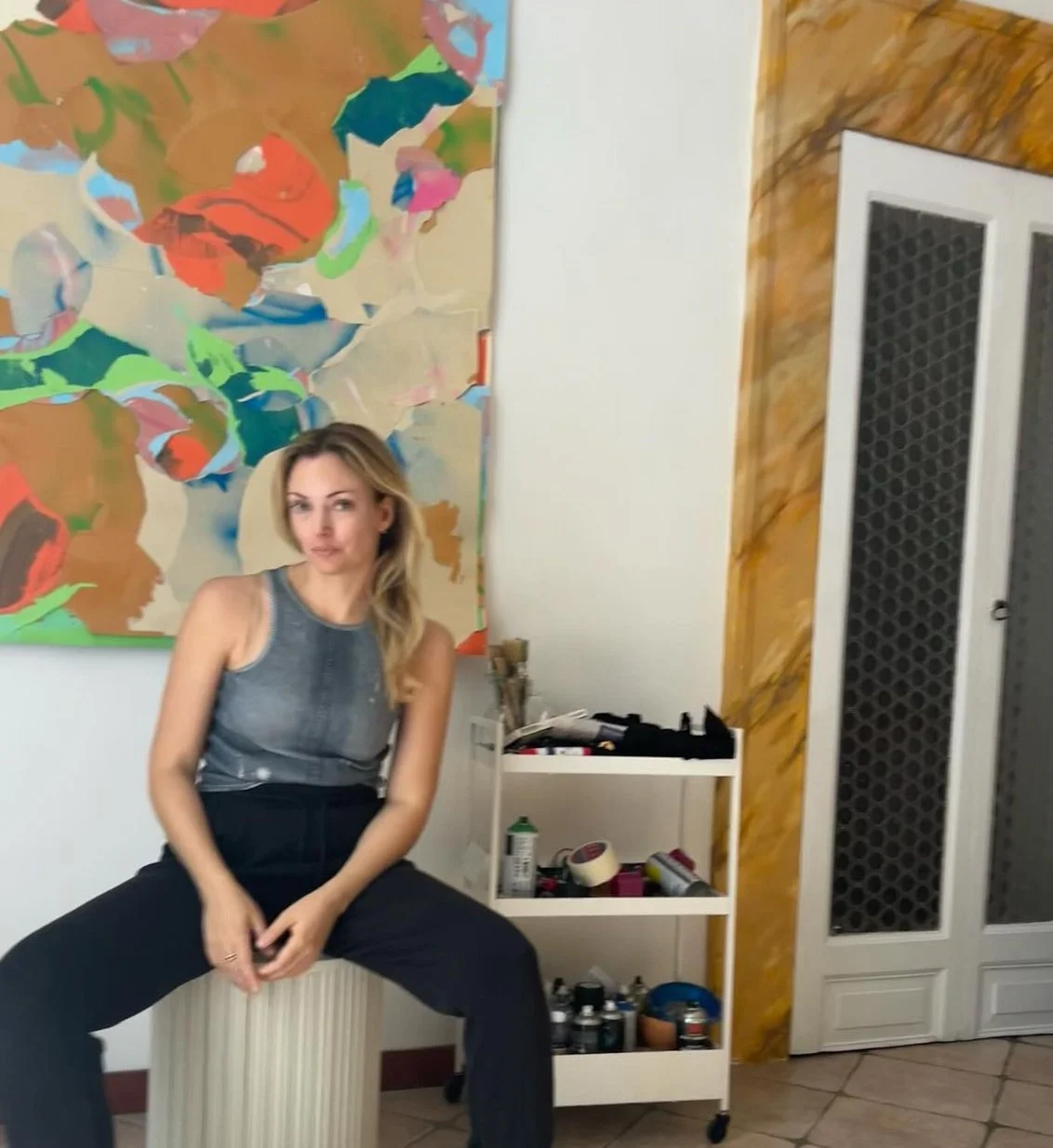 A young woman sitting on a white base drum in a room with abstract artwork on the wall. She is wearing a sleeveless grey top and black pants, with her hands resting on her lap. There is a small white shelf with art supplies and spray paint cans behind her, and a door with a patterned screen on the right side.
