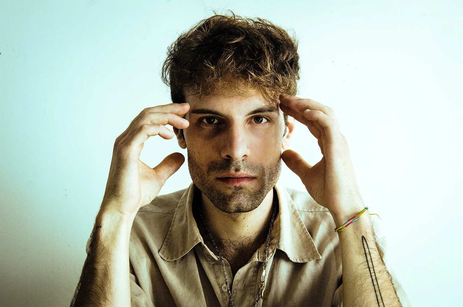 A young man with brown, curly hair, light stubble, wearing a beige shirt, holding his hands near his head, looking directly at the camera against a plain light background.