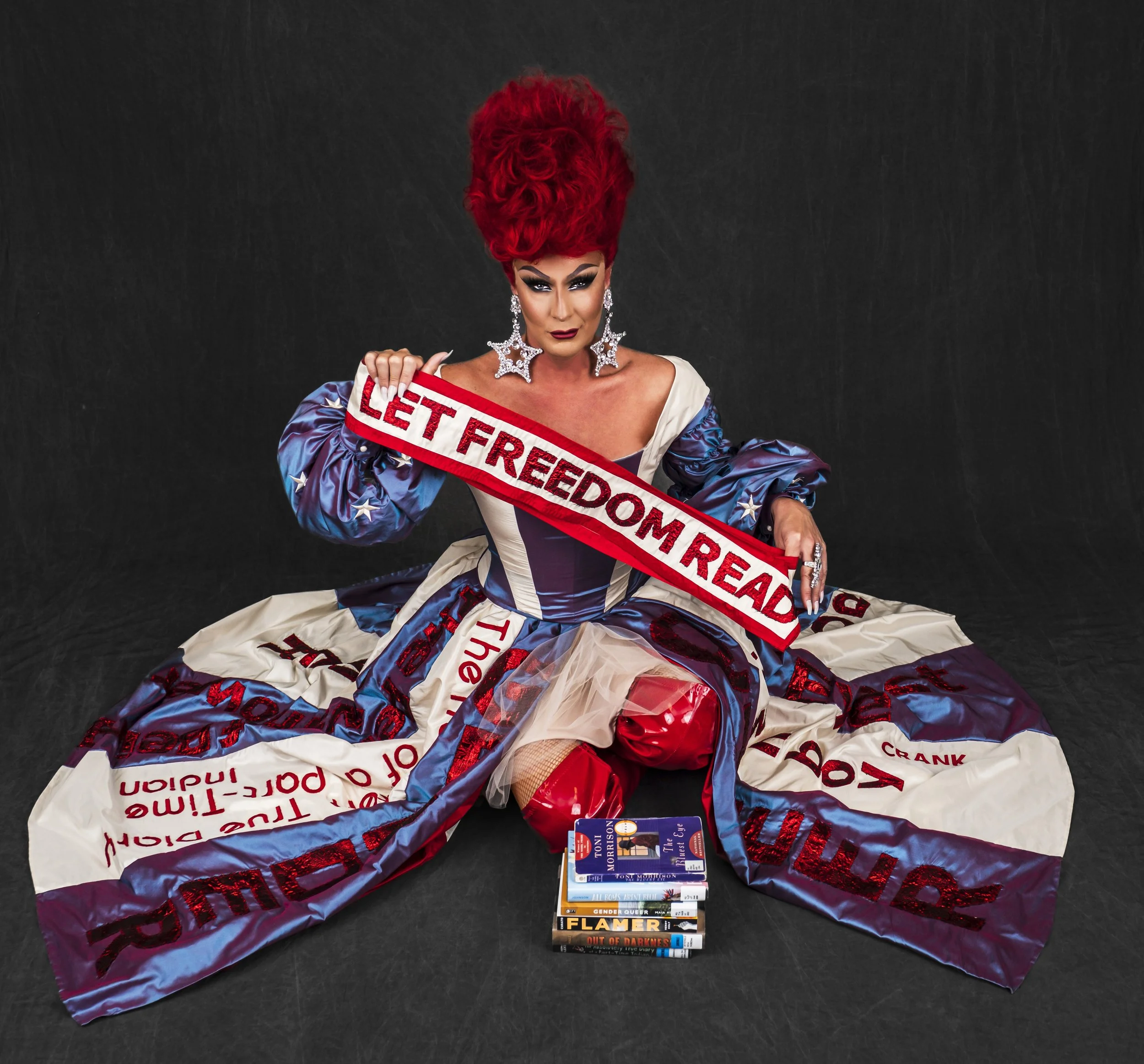 A drag queen holding a sash that reads 'Let Freedom Read'. They have vibrant red hair styled tall and dramatic makeup. The person is sitting on a black background with a stack of banned books placed on the floor in front of them.