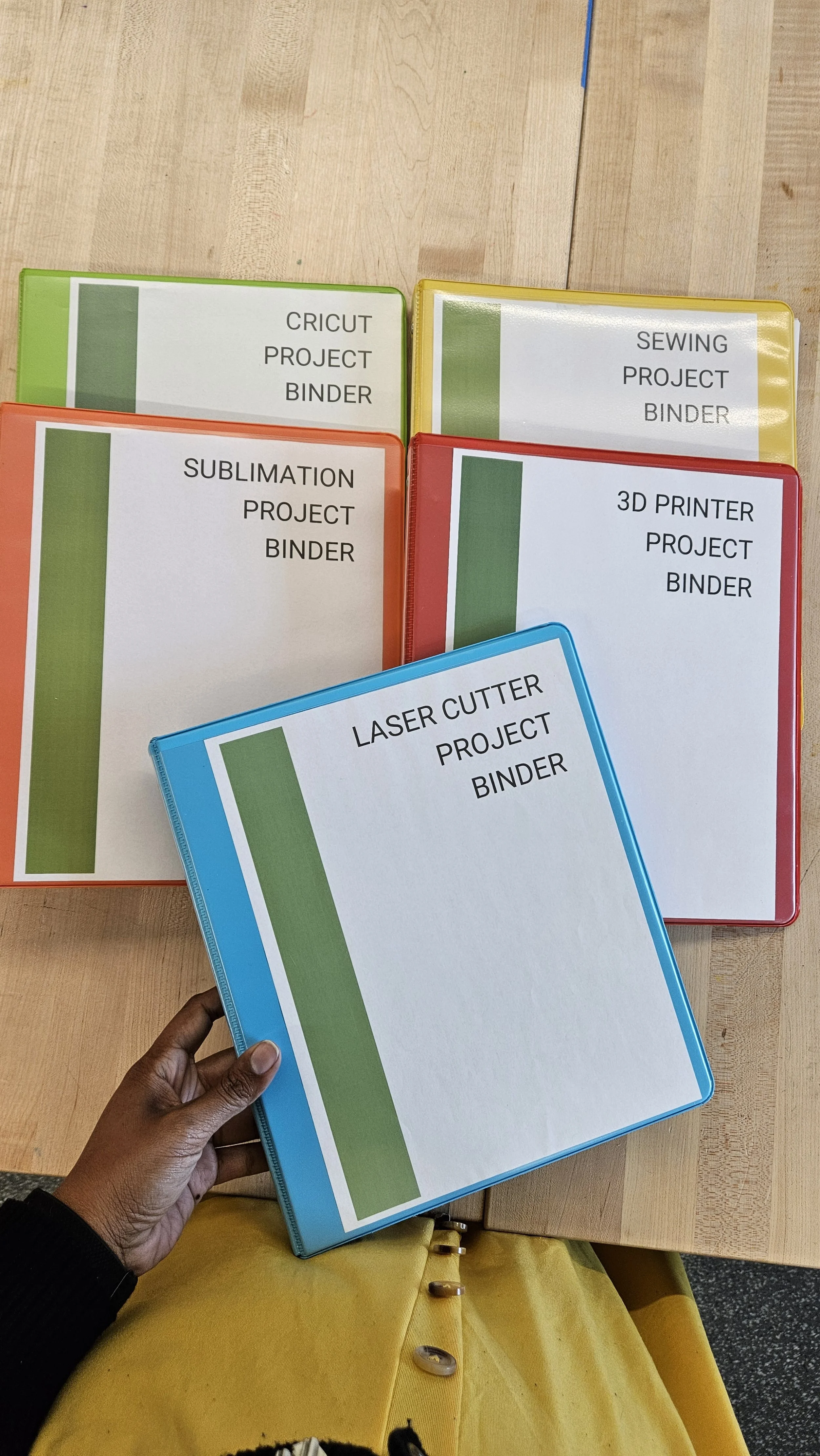 Five project binders labeled for Cricut, Sewing, Sublimation, 3D Printer, and Laser Cutter, on a wooden table.