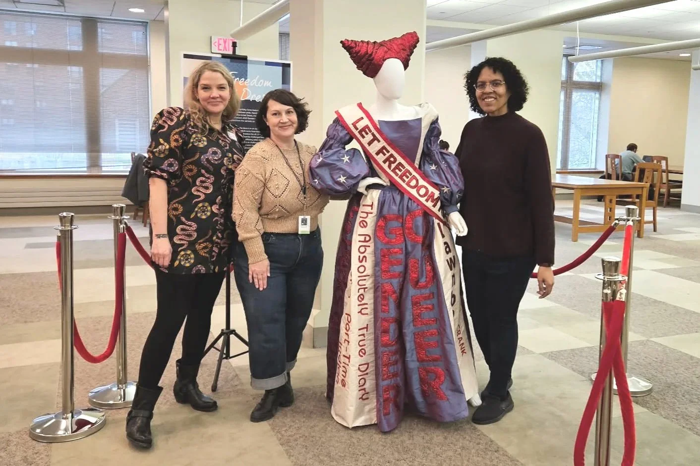 Three women standing next to a mannequin dressed in a costume with a sash that reads 'LET FREEDOM READ.' The women are smiling and posing for the photo in a room with large windows and a red velvet rope barrier.