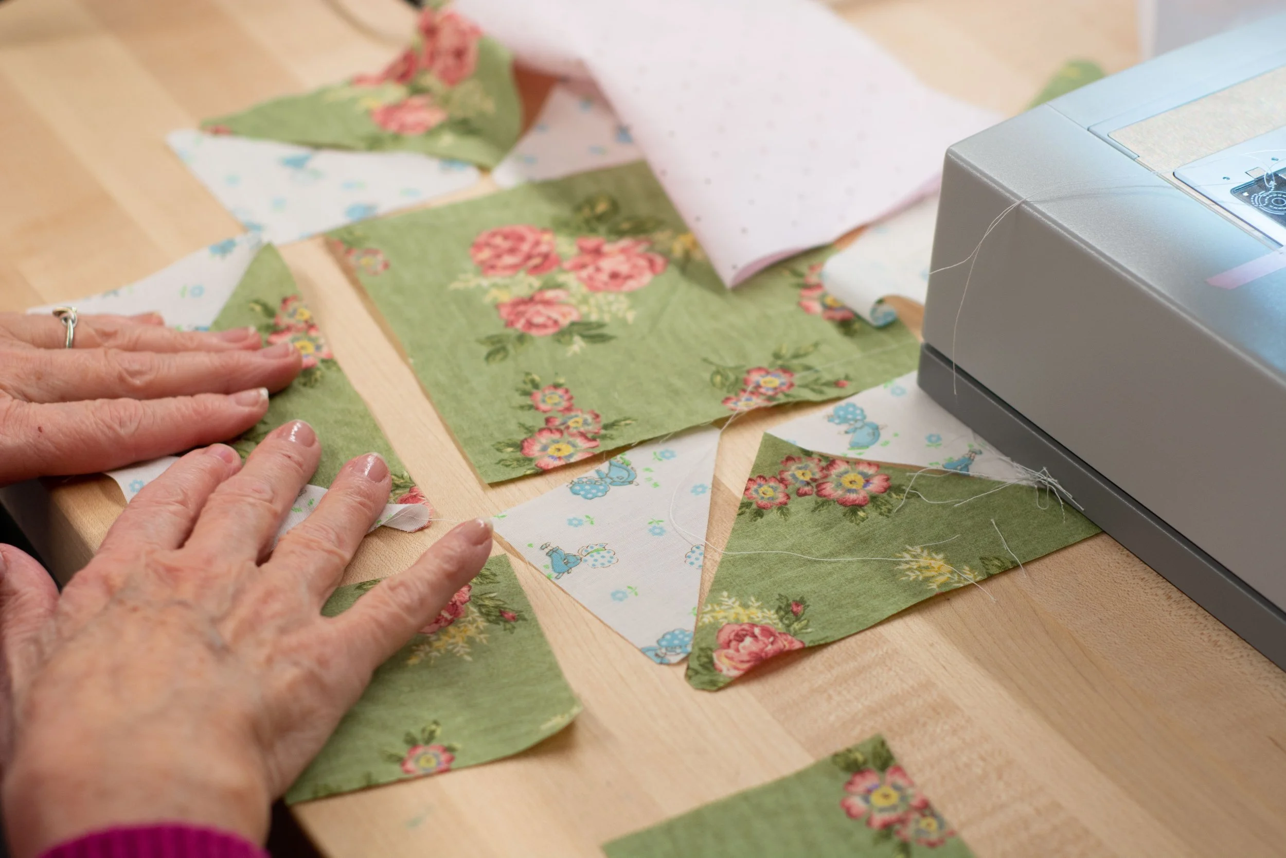 Someone sewing a quilt with floral and patterned fabric pieces on a wooden table.