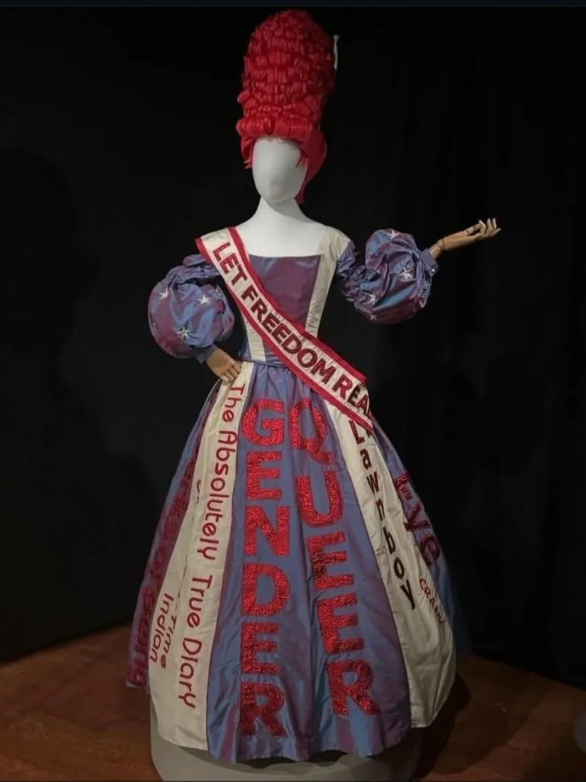 A mannequin wearing the traveling "Let Freedom Read" dress, with a large red hairdo. The dress has messages about freedom, liberty, and activism embroidered on it.