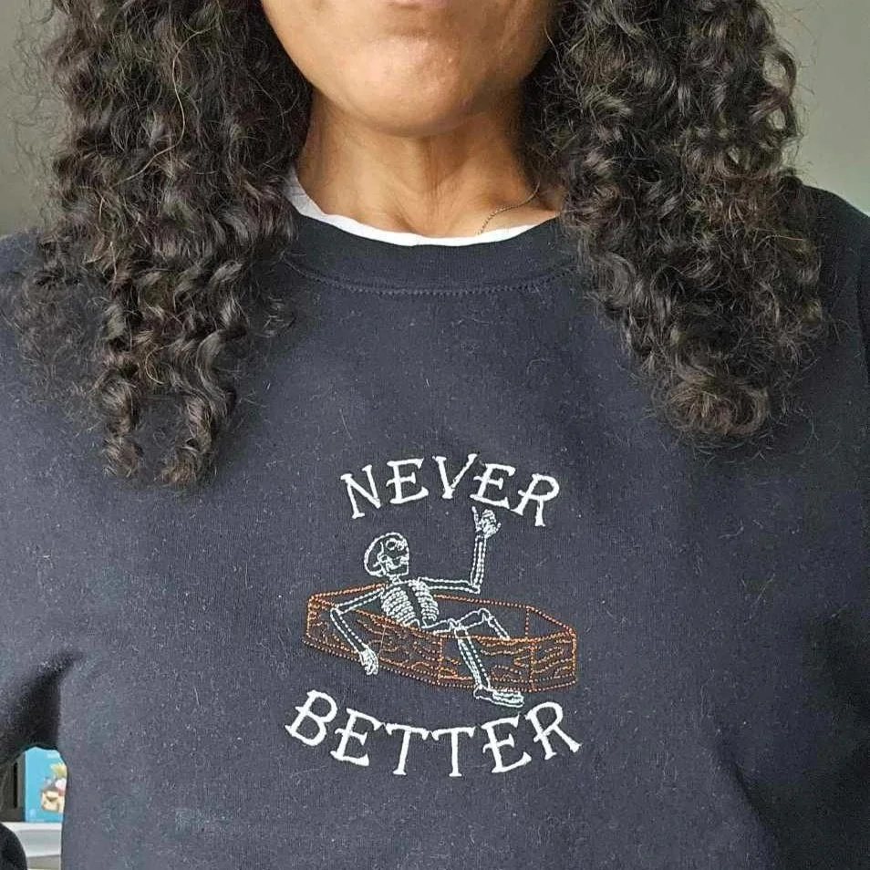 A person wearing a black sweatshirt with an embroidered design of a skeleton lying in a coffin and the text "Never Better."