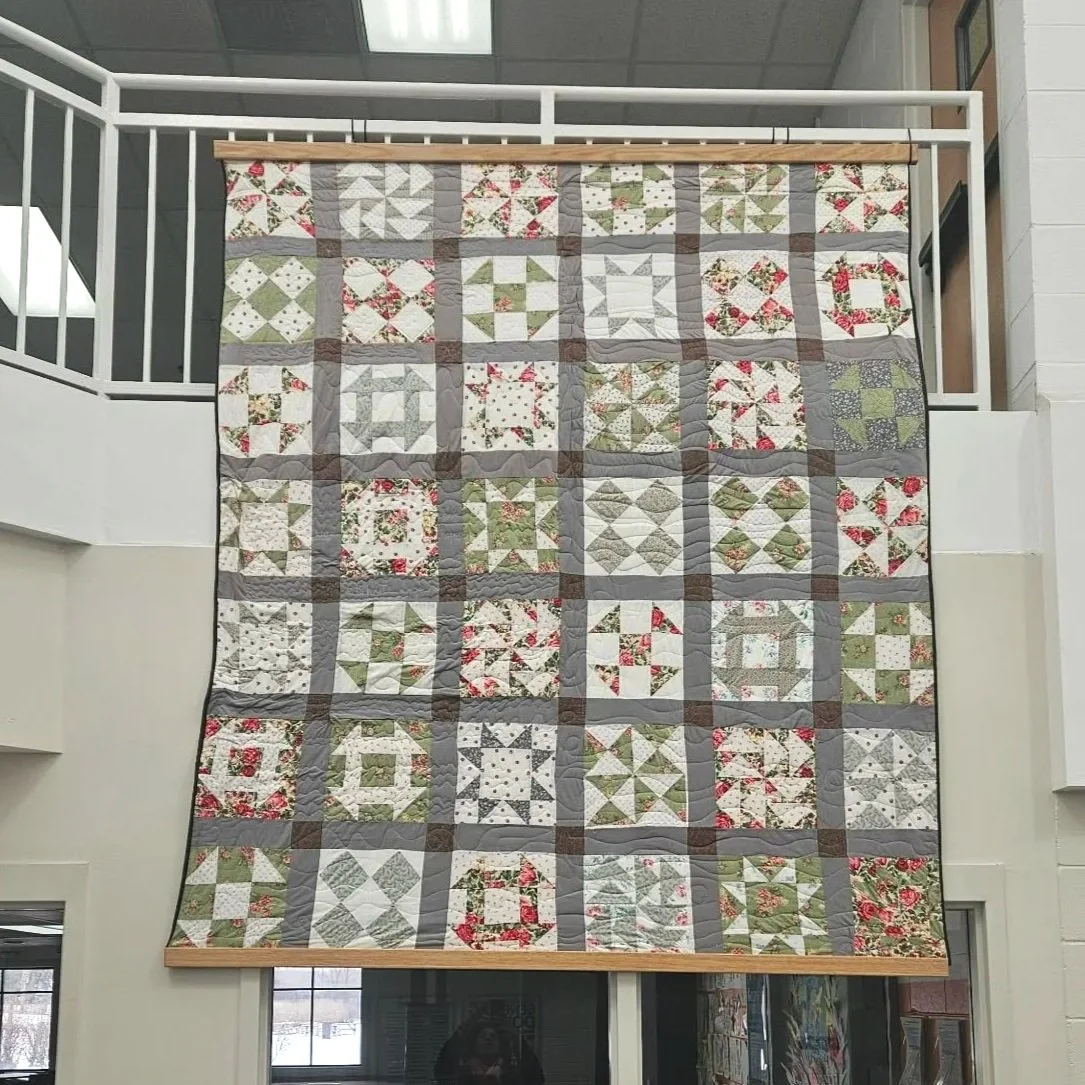 Large quilt with a patchwork pattern of various designs in white, green, red, and gray, hanging on a wooden rod in a room with staircases and windows.