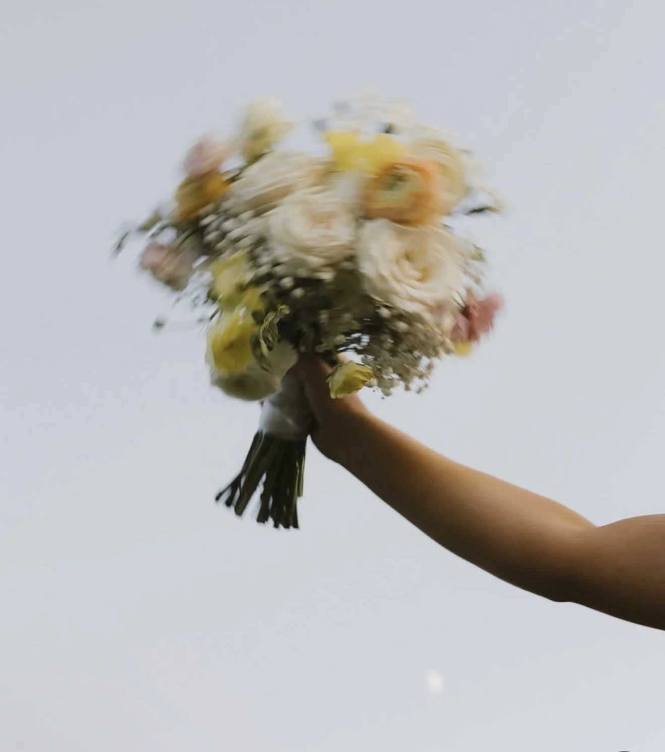 A person holding a bouquet of white, yellow, orange, and pink flowers against a pale sky background.