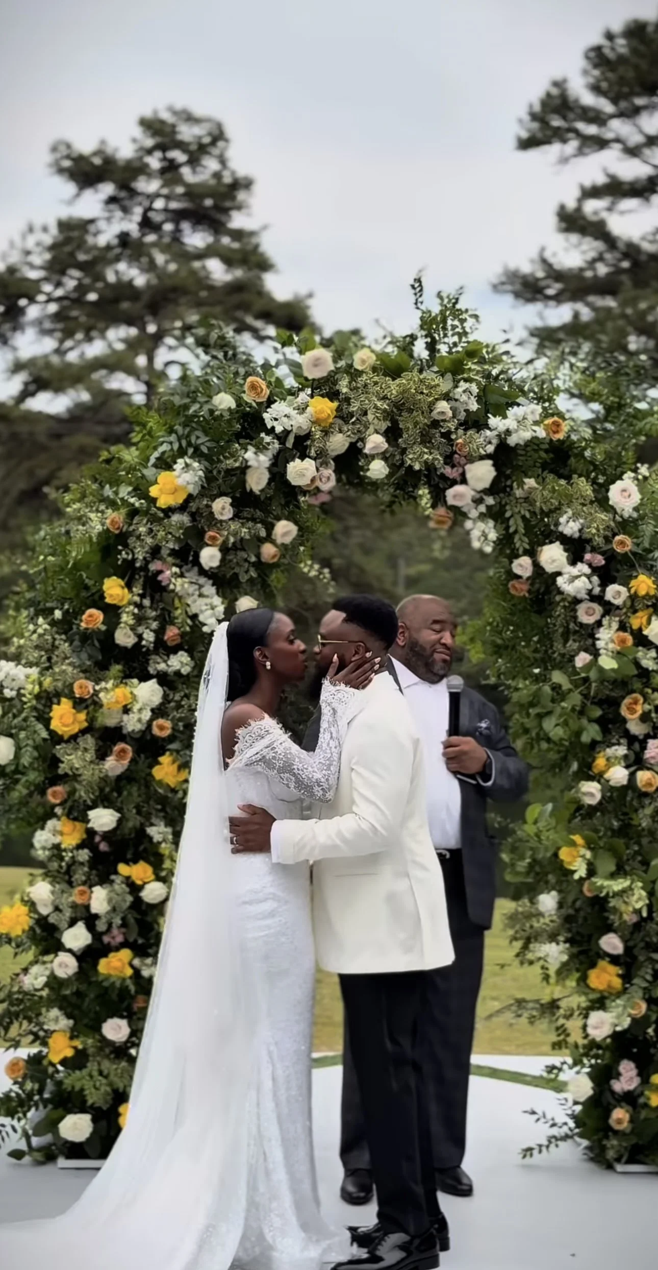 A wedding ceremony with a bride and groom standing under a floral arch, with an officiant holding a microphone nearby.