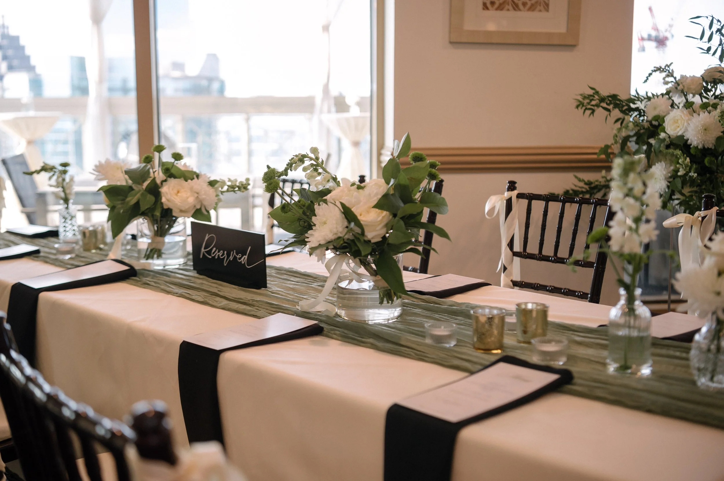 A decorated table for a wedding or special event, with white flowers in glass vases, a reserved sign, candle holders, and black napkins on a cream tablecloth with a green table runner.
