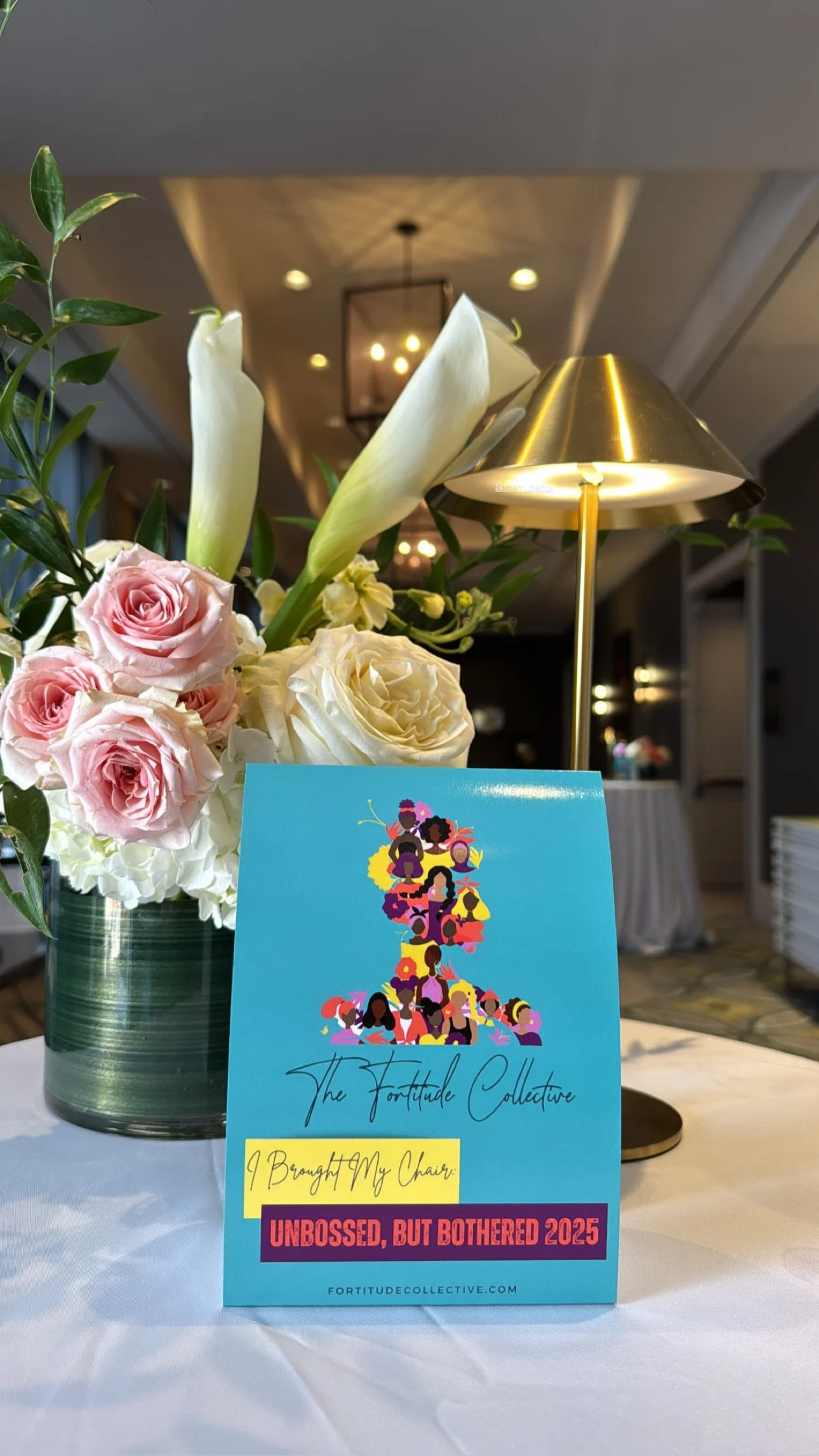 A colorful event card with a silhouette of diverse women made of illustrations, placed on a table next to a floral bouquet with pink roses and white flowers, and a gold lamp in the background.