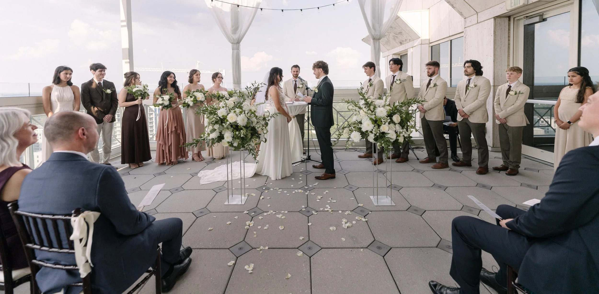 A wedding ceremony taking place on a rooftop with a city skyline in the background. The bride and groom are standing at the altar, exchanging vows, surrounded by bridesmaids and groomsmen. Floral arrangements flank the couple, and guests seated in th