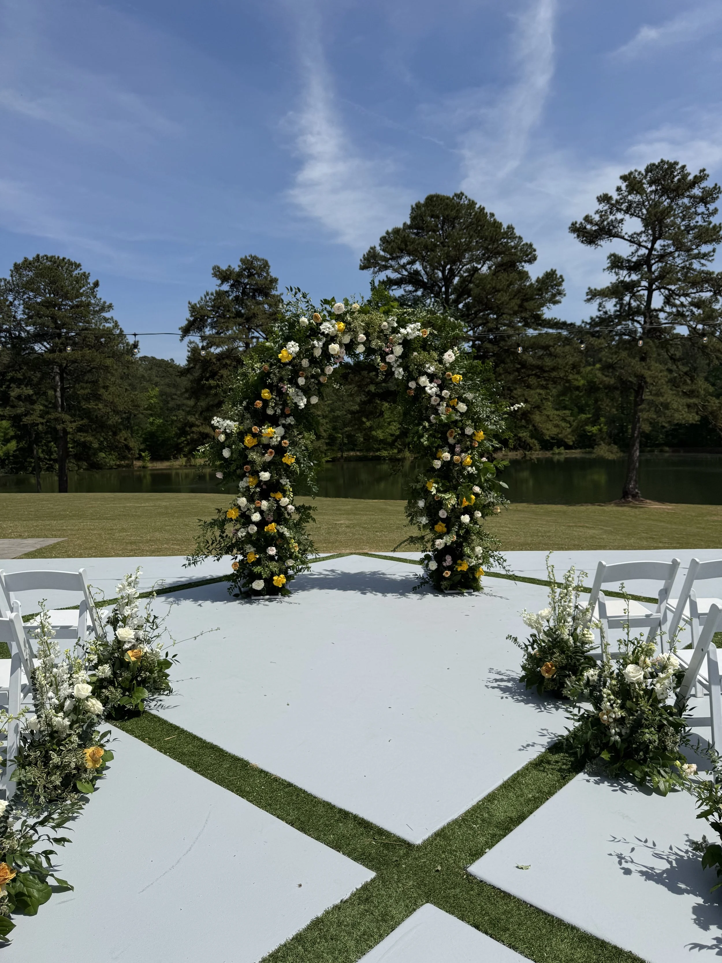 Outdoor wedding ceremony setup with a white aisle, floral arch, and chairs on both sides, near a lake with trees and a blue sky in the background.