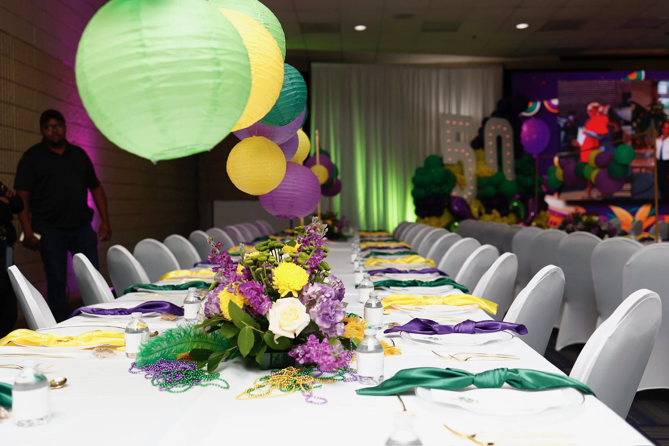 Decorated banquet table with colorful paper lanterns hanging above, floral centerpieces, and chairs with yellow, purple, and green sashes, set for a 50th birthday celebration.