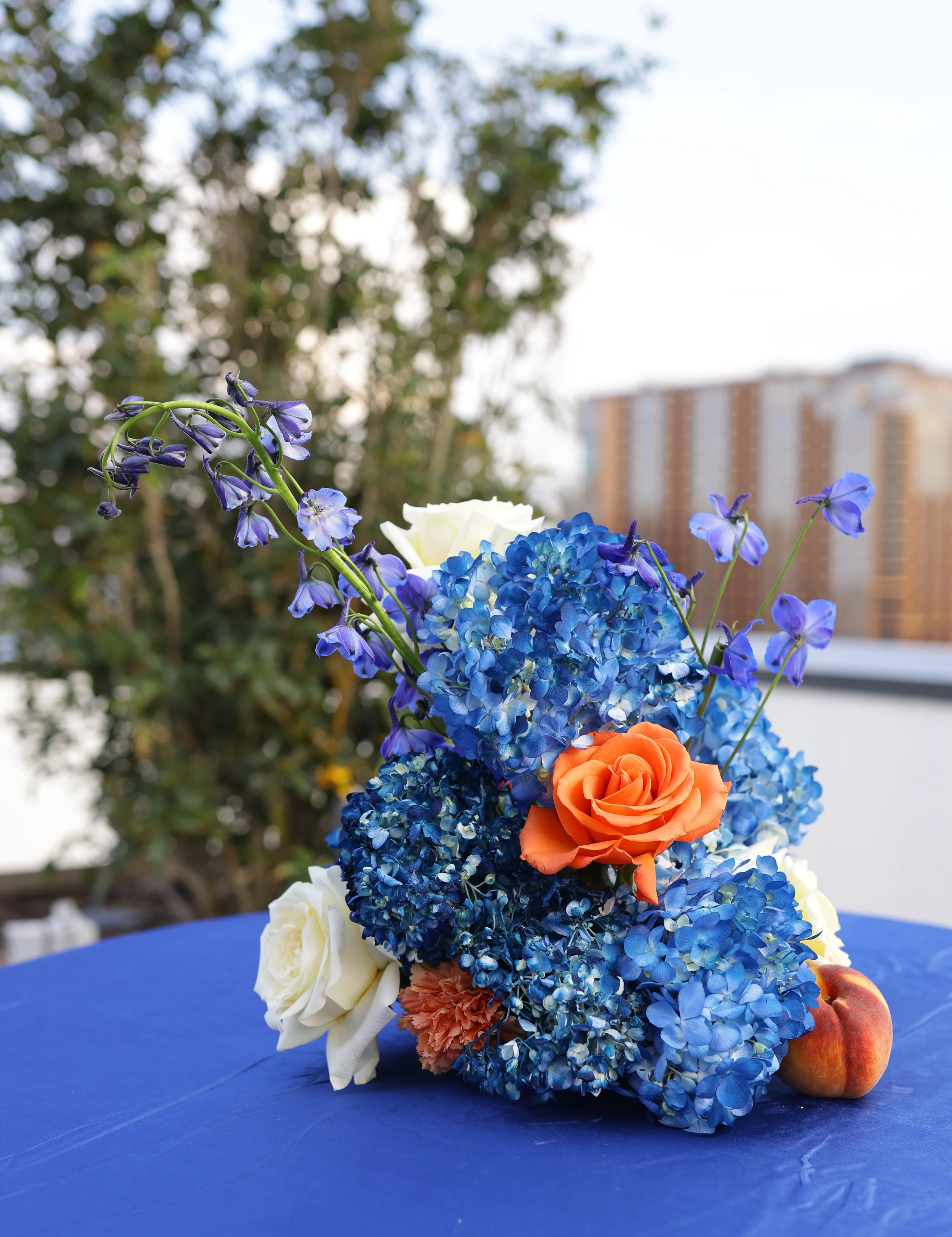 Colorful flower arrangement on a blue table with a blurred outdoor background of trees and buildings.