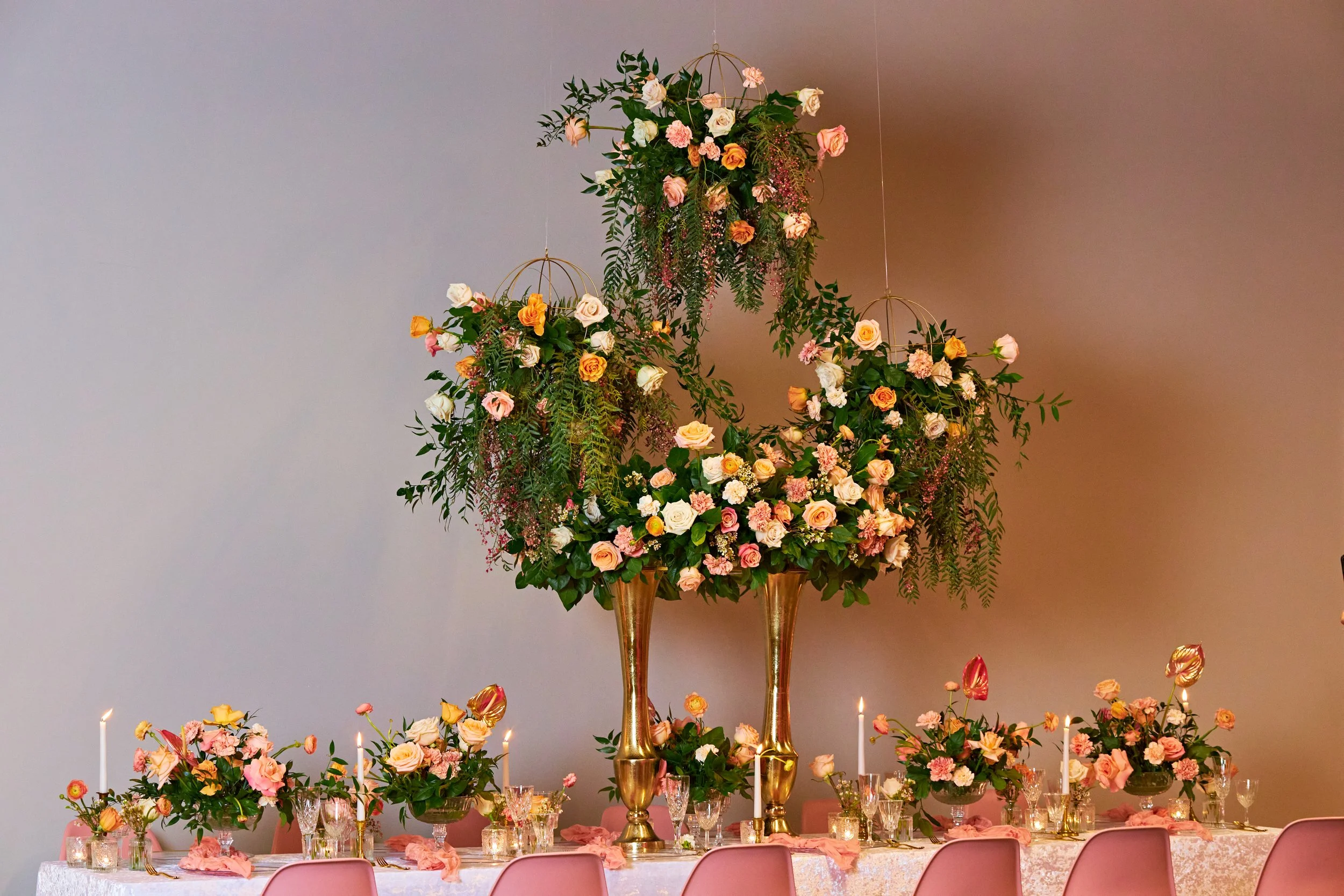 A decorated banquet table with floral arrangements, candles, and pink chairs, featuring a large floral centerpiece with white, pink, and peach roses and greenery.