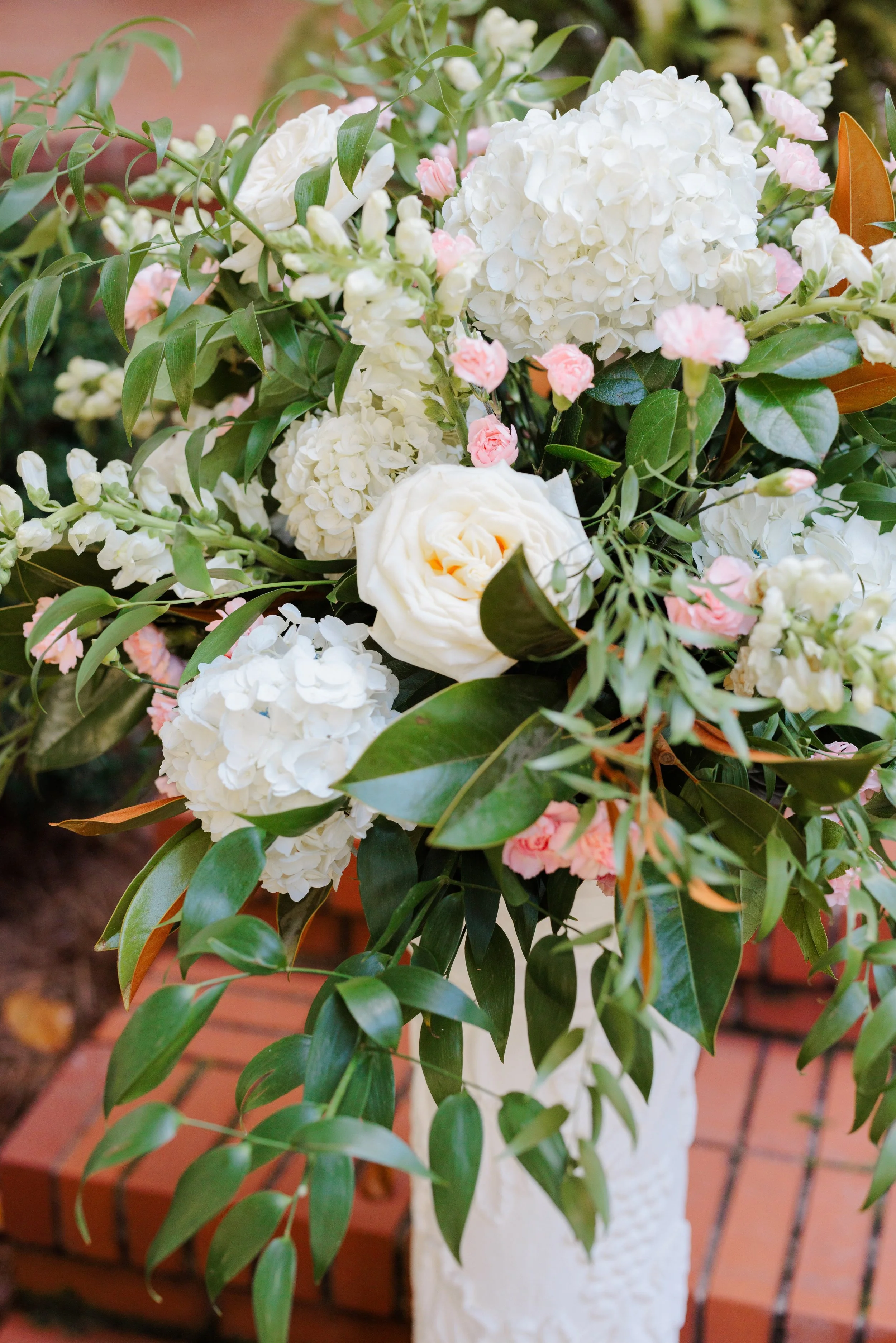 A floral arrangement with white hydrangeas, roses, and pink carnations in a white vase on a brick surface.