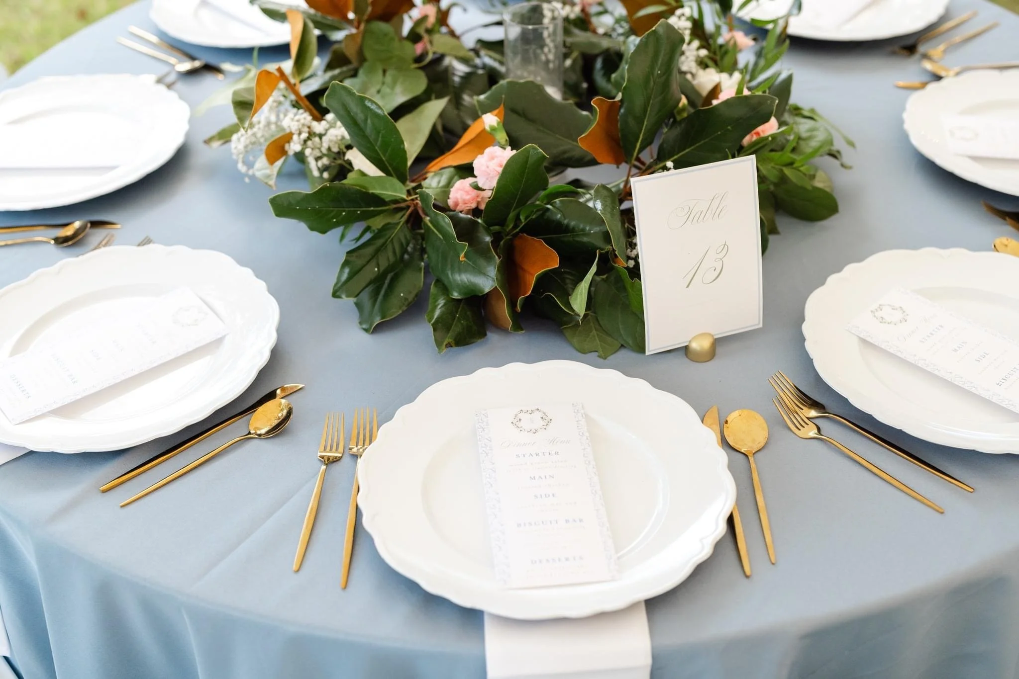 Elegant wedding table setting with white plates, gold flatware, a gray tablecloth, a floral centerpiece with green leaves, baby's breath, and pink flowers, and a table card labeled "Table 13".