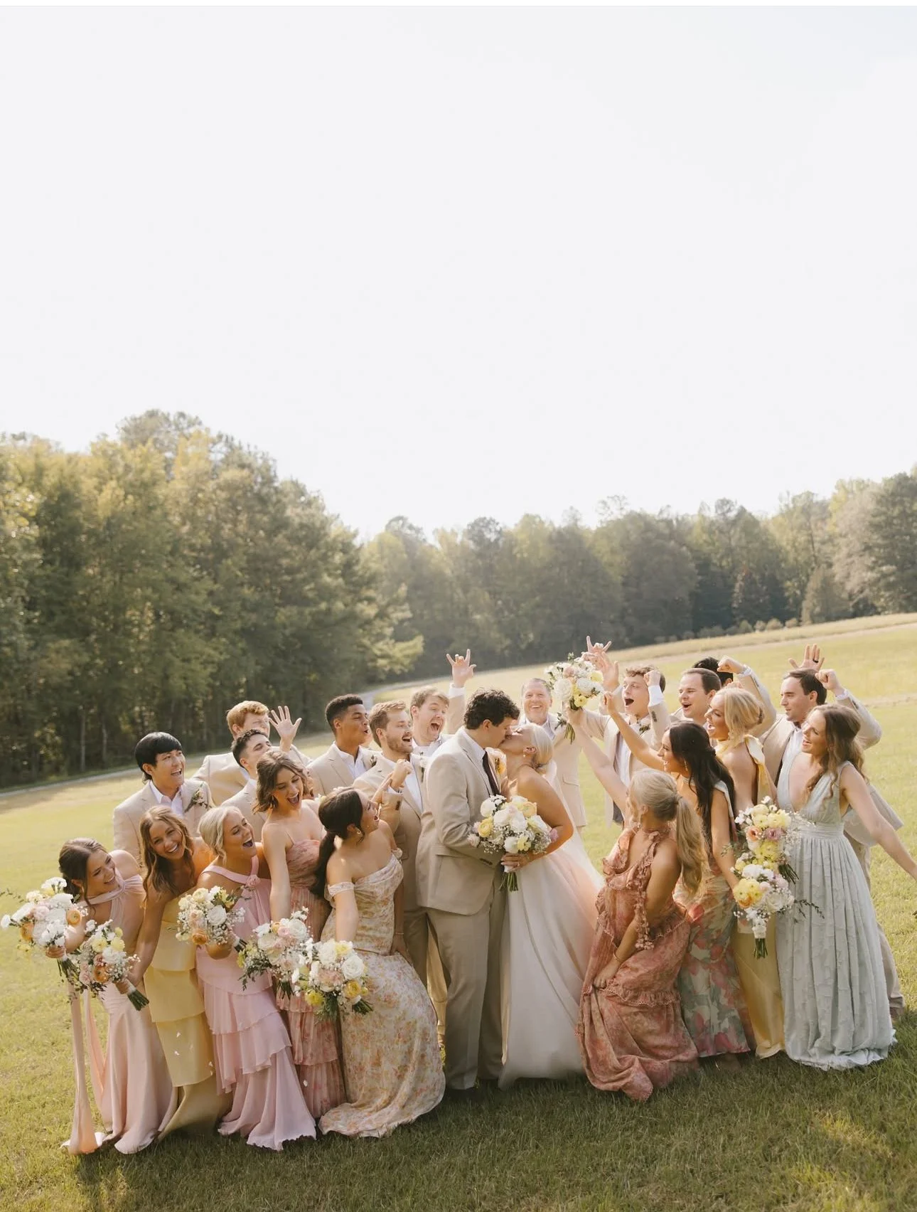 A large group of wedding guests gathered outdoors on a grassy field, celebrating a wedding. The bride and groom are in the center, kissing, with the bride holding a bouquet. The group is smiling, cheering, and holding bouquets, with a backdrop of tre