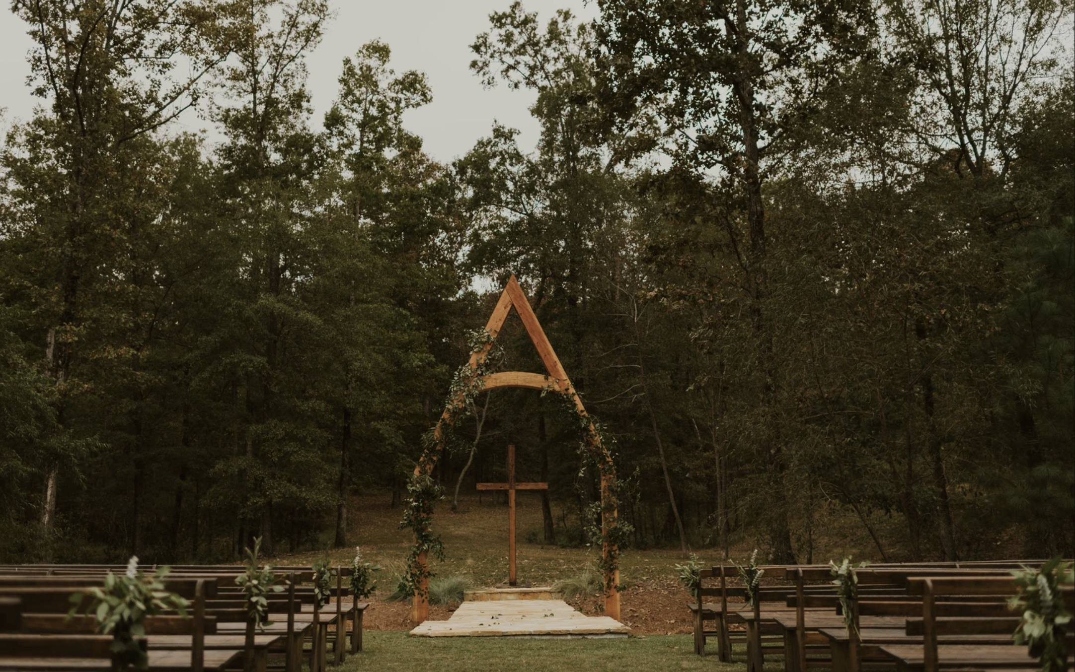 Outdoor wedding ceremony setup with wooden arch decorated with greenery and flowers, overlooking forest, with rows of benches on either side.