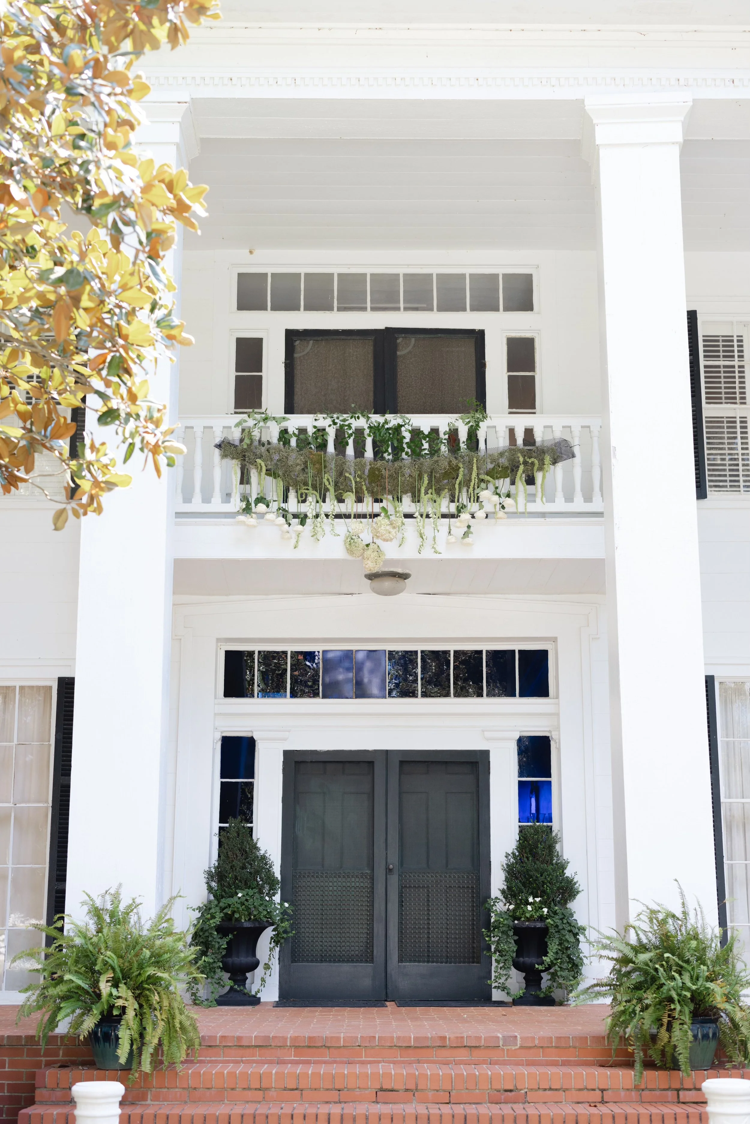 White multi-story house with black double doors, hanging plants on balcony, potted plants on brick steps, shaded front porch, and white walls.