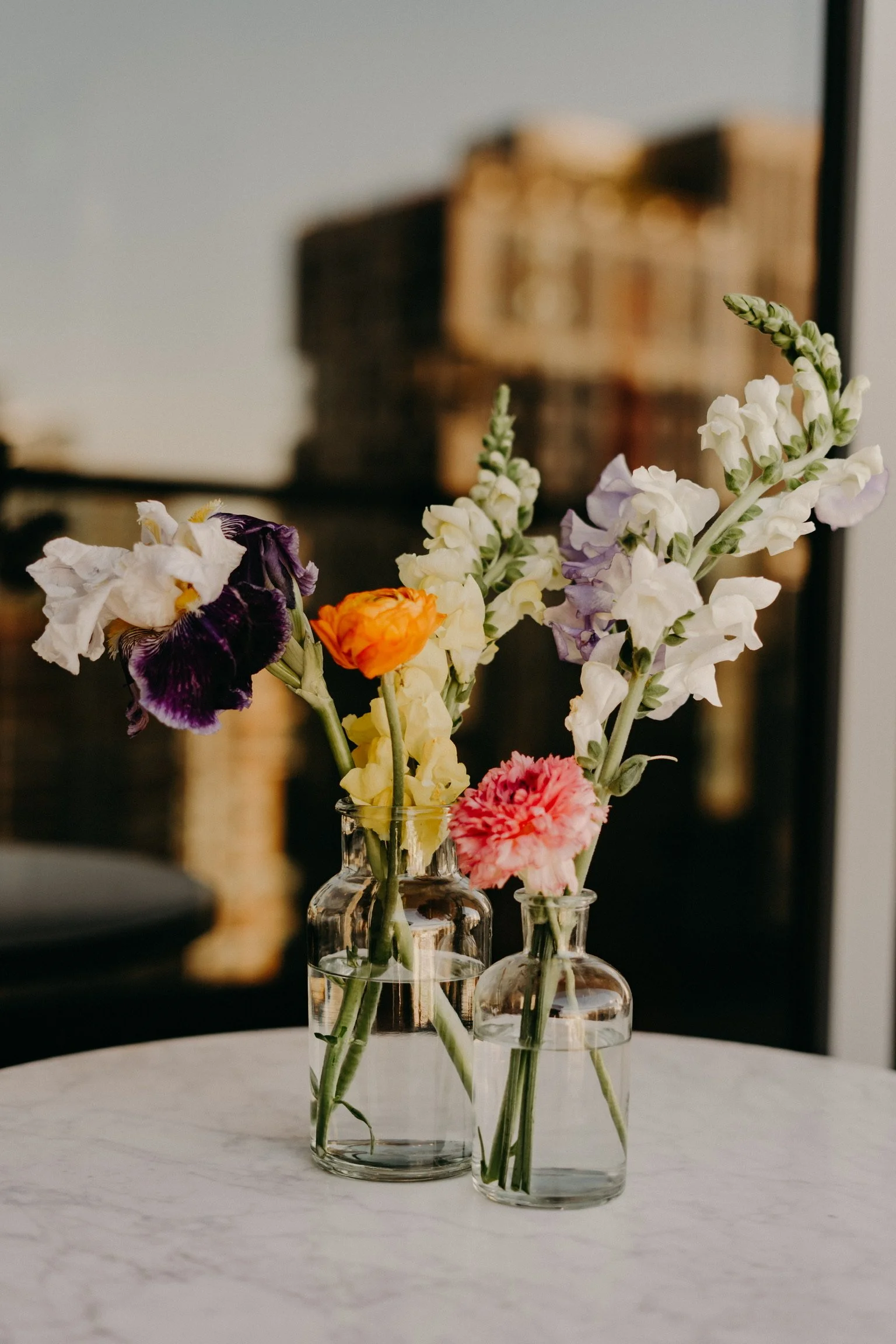 Two glass vases filled with water and colorful flowers, set on a white marble table, with blurred cityscape background.