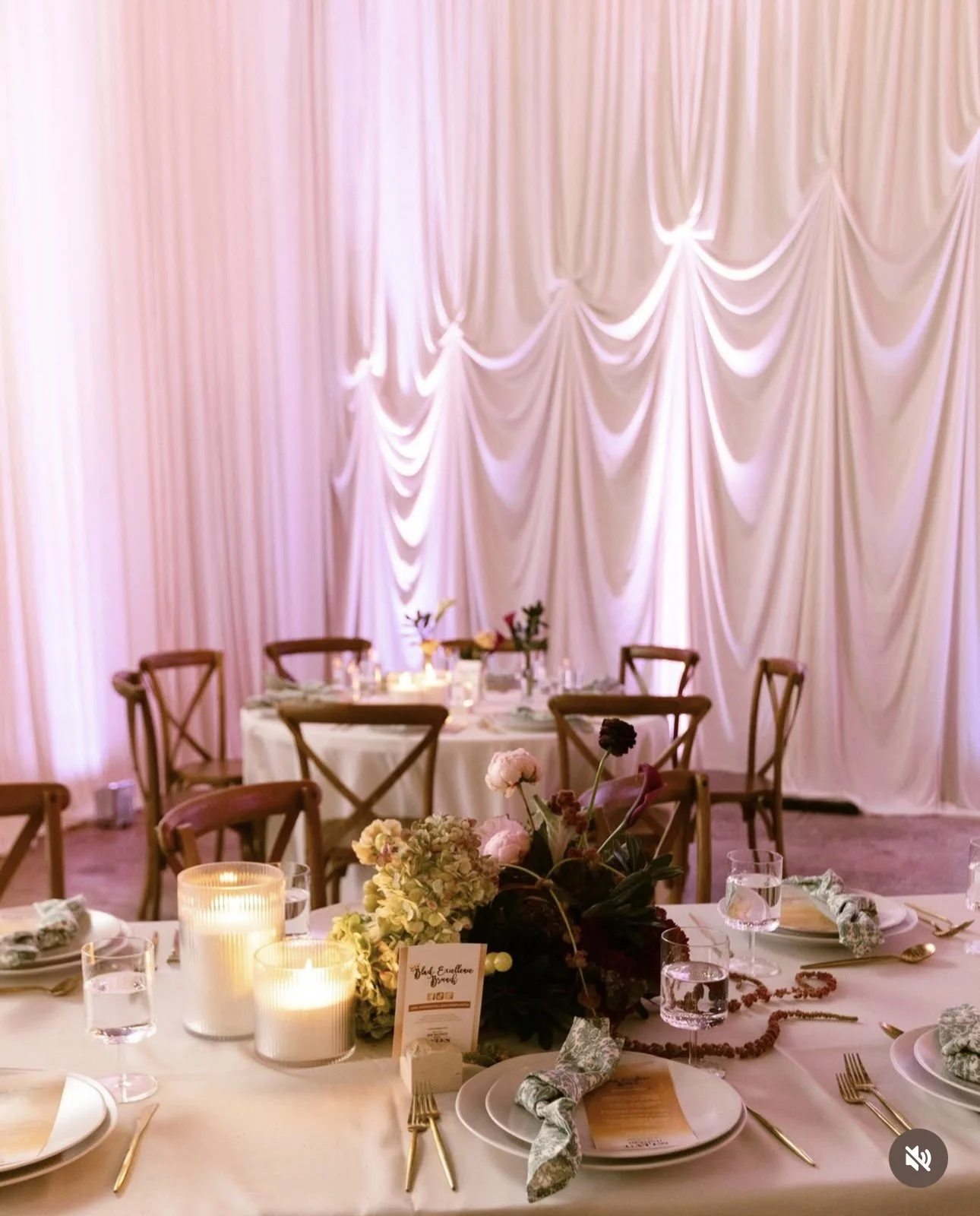 Elegant dining table set with candles, flowers, and candles against a pink, draped backdrop for a celebration or wedding reception.
