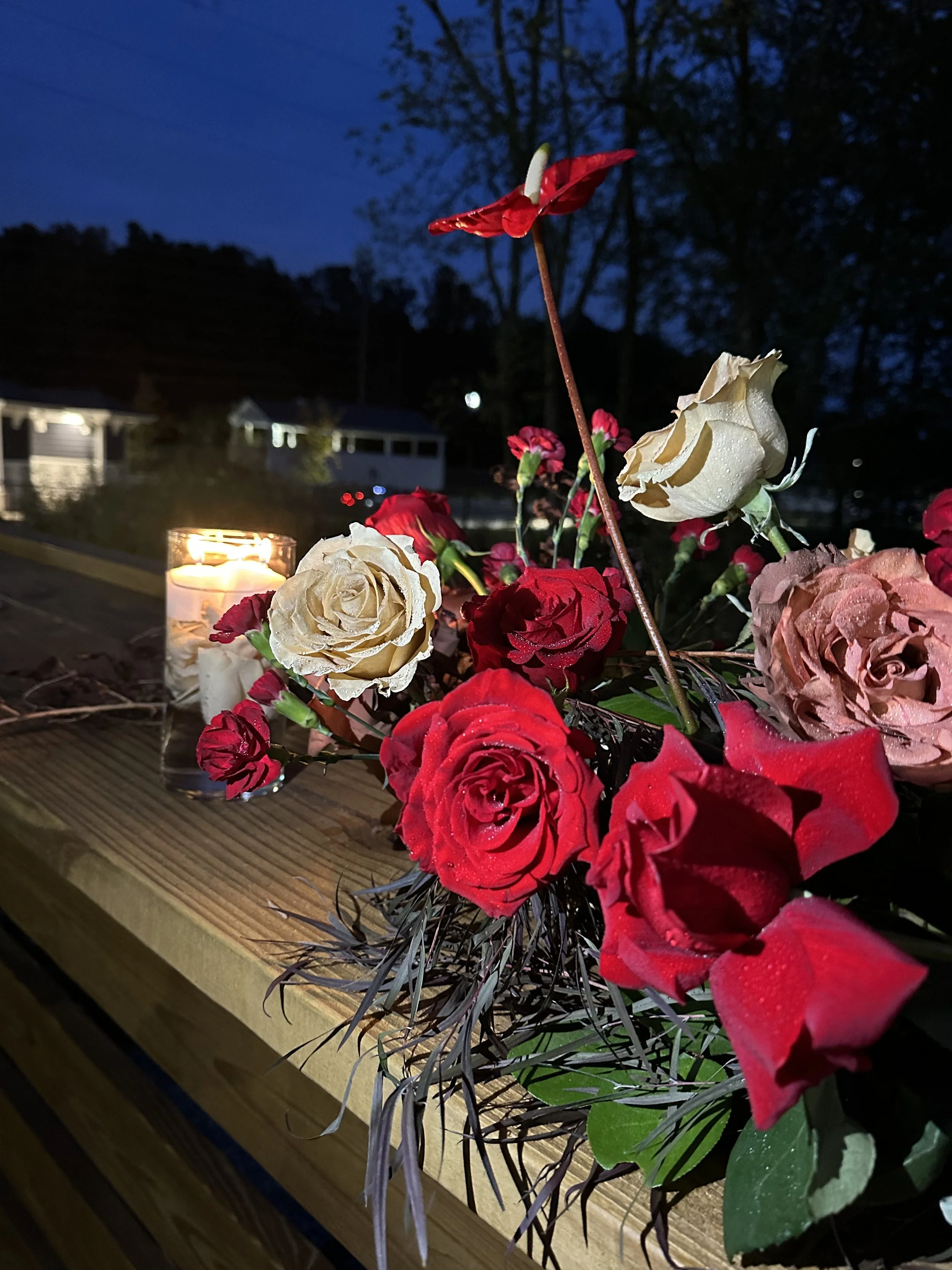 A floral arrangement with red and white roses and pink flowers, lit by a candle, on a wooden surface outdoors at night, with trees and houses in the background.