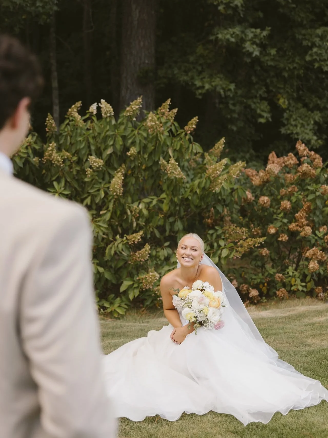 A bride in a strapless white wedding gown sitting on the grass, holding a bouquet of white and pale pink flowers, smiling at a groom in a beige suit who is partly visible in the foreground.