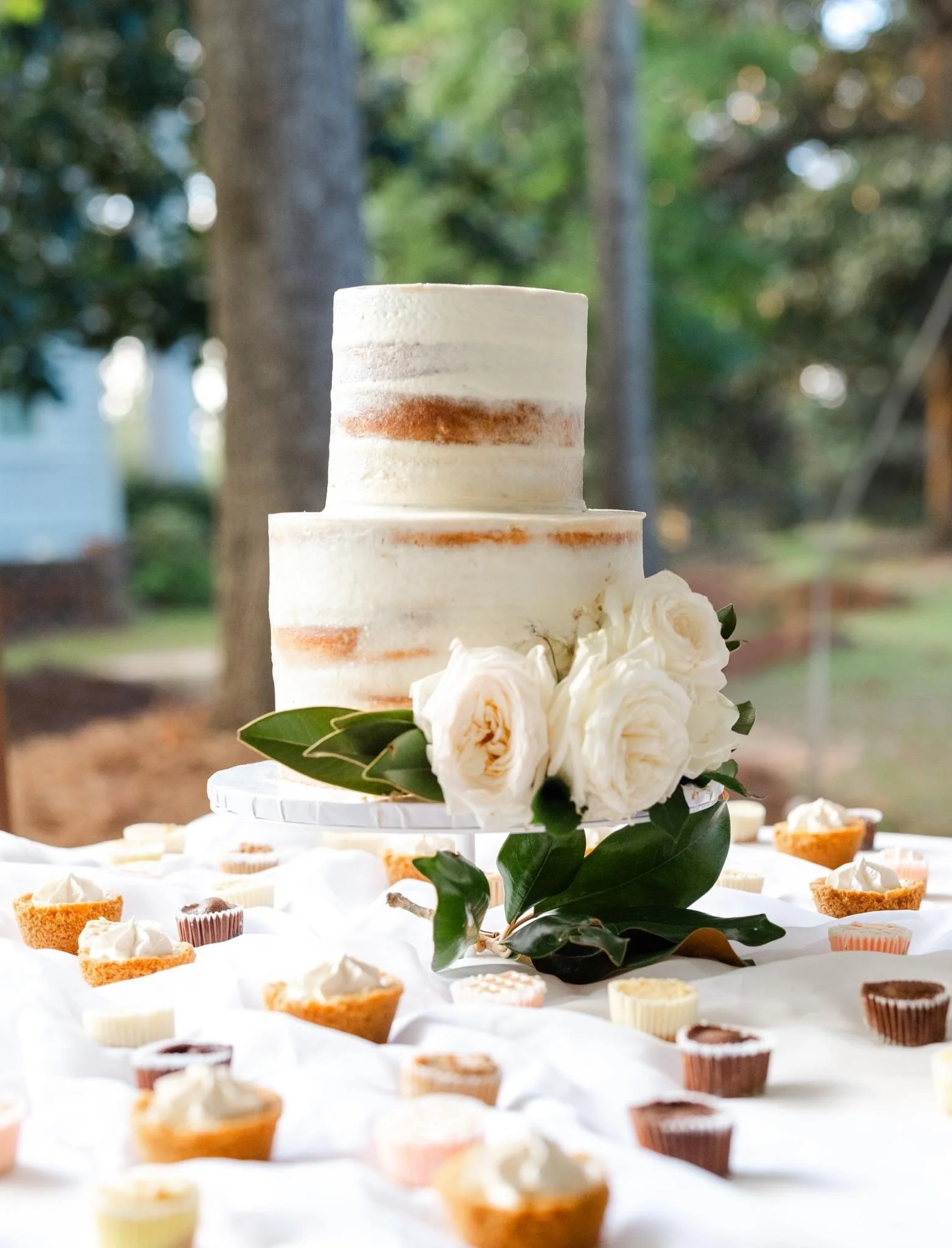 A two-tiered wedding cake with white frosting and a semi-naked design, decorated with white roses and green leaves, surrounded by small cupcakes on a table outdoors.