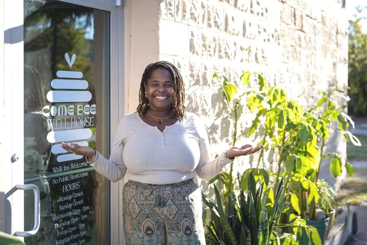 A woman with dreadlocks smiling and standing outdoors next to a glass door with a sign, surrounded by green plants, in front of a stone wall.