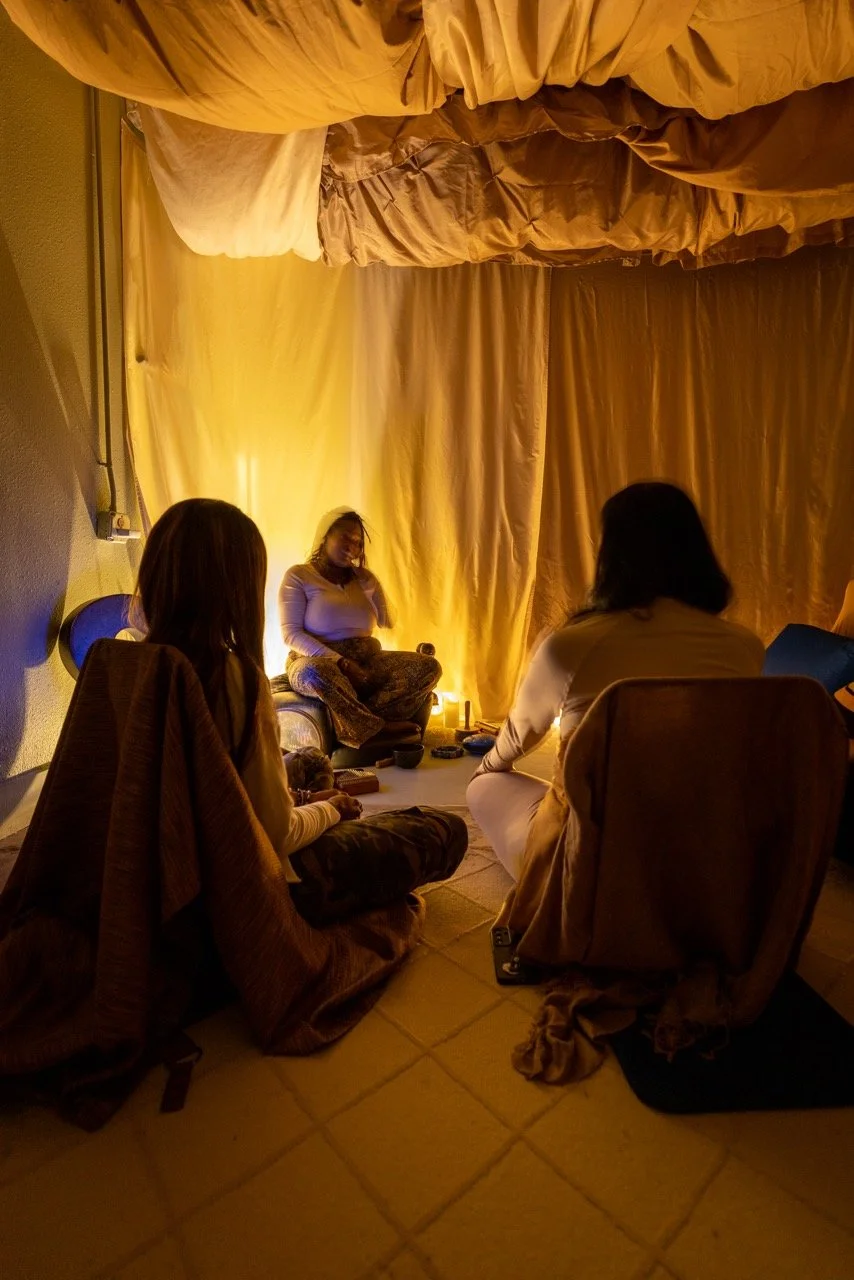 Three women sitting on the floor in a dimly lit room, engaged in conversation or meditation, with a woman seated on a cushion against a golden curtain backdrop, illuminated by a candle.