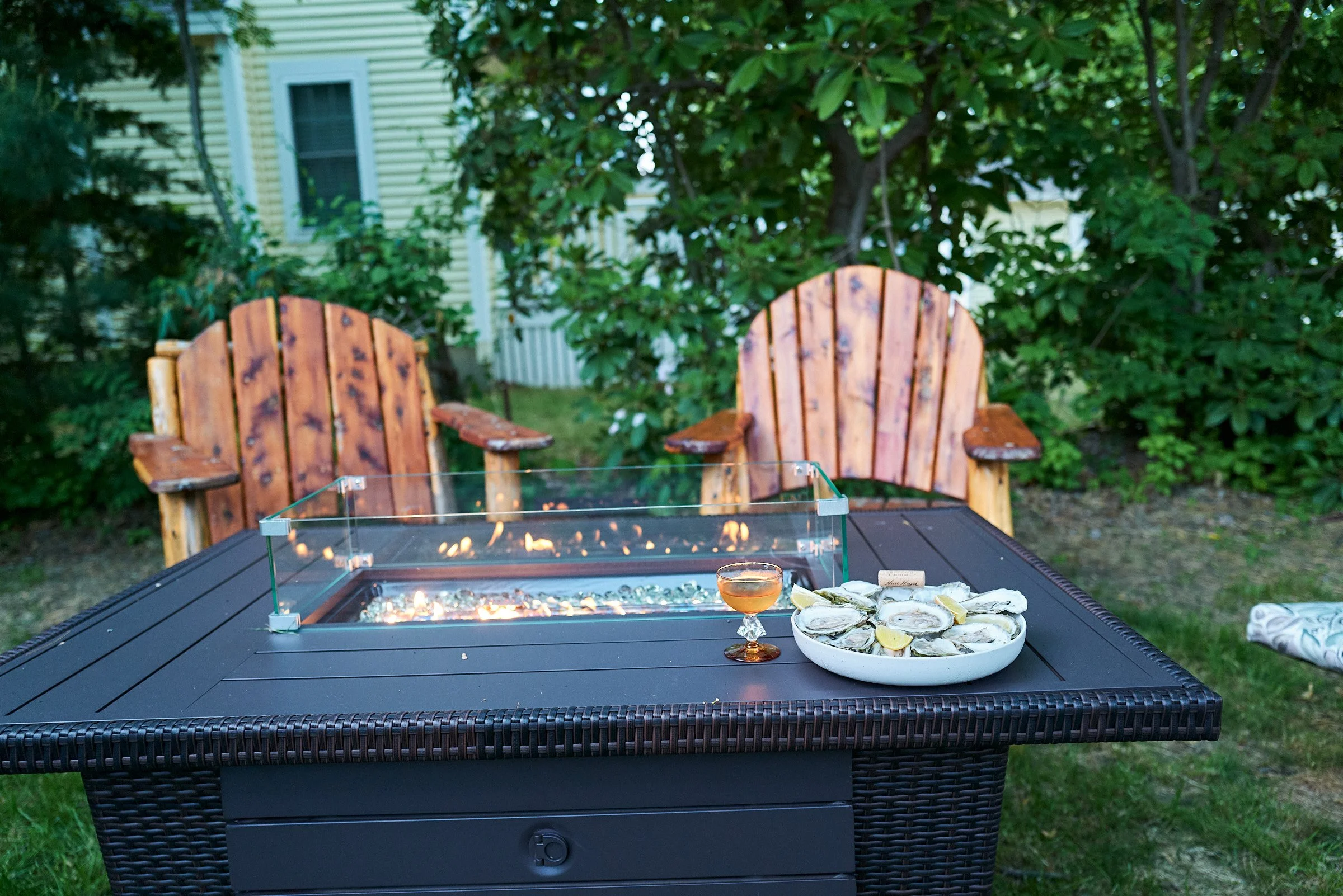 Outdoor patio with a fire pit table, two wooden Adirondack chairs, a plate of oysters with lemon wedges, and a glass of wine.
