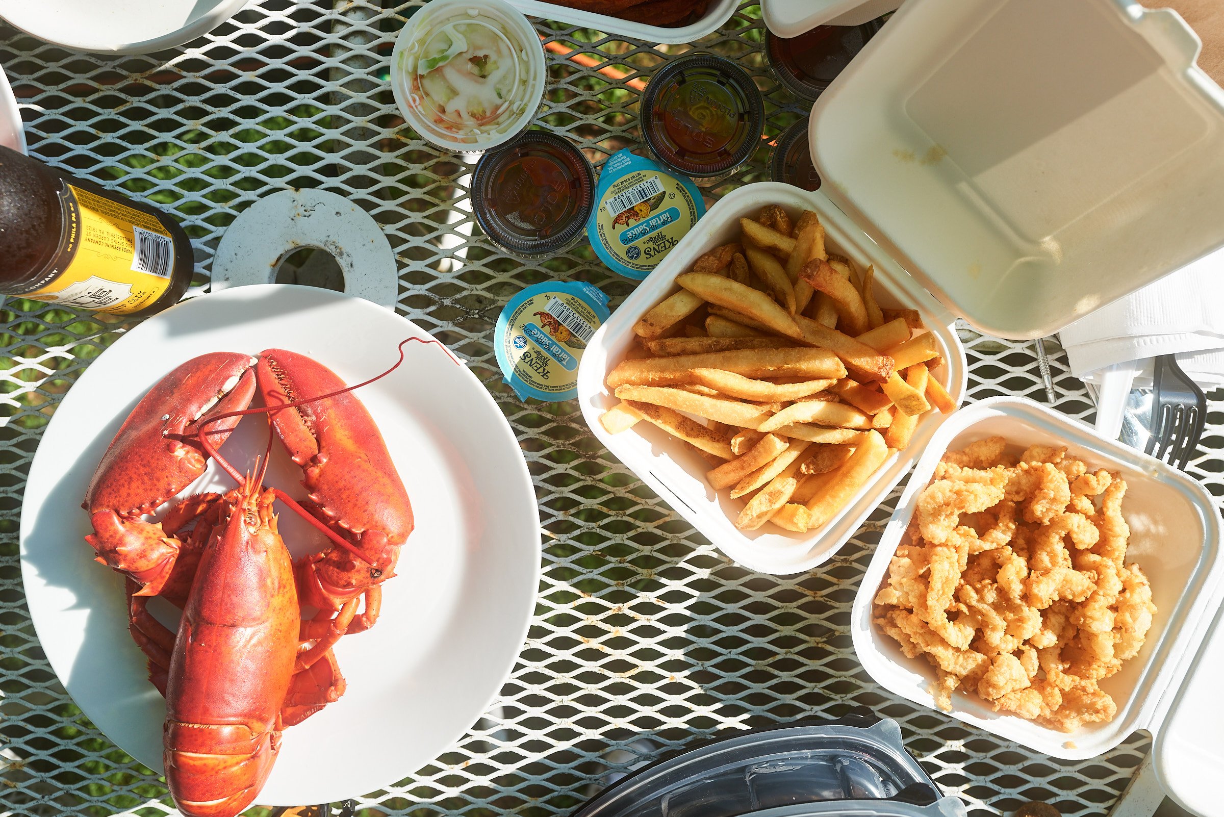 Top view of a picnic table with cooked lobster on a white plate, French fries, fried seafood, coleslaw, dipping sauces, and a bottle of beverage.