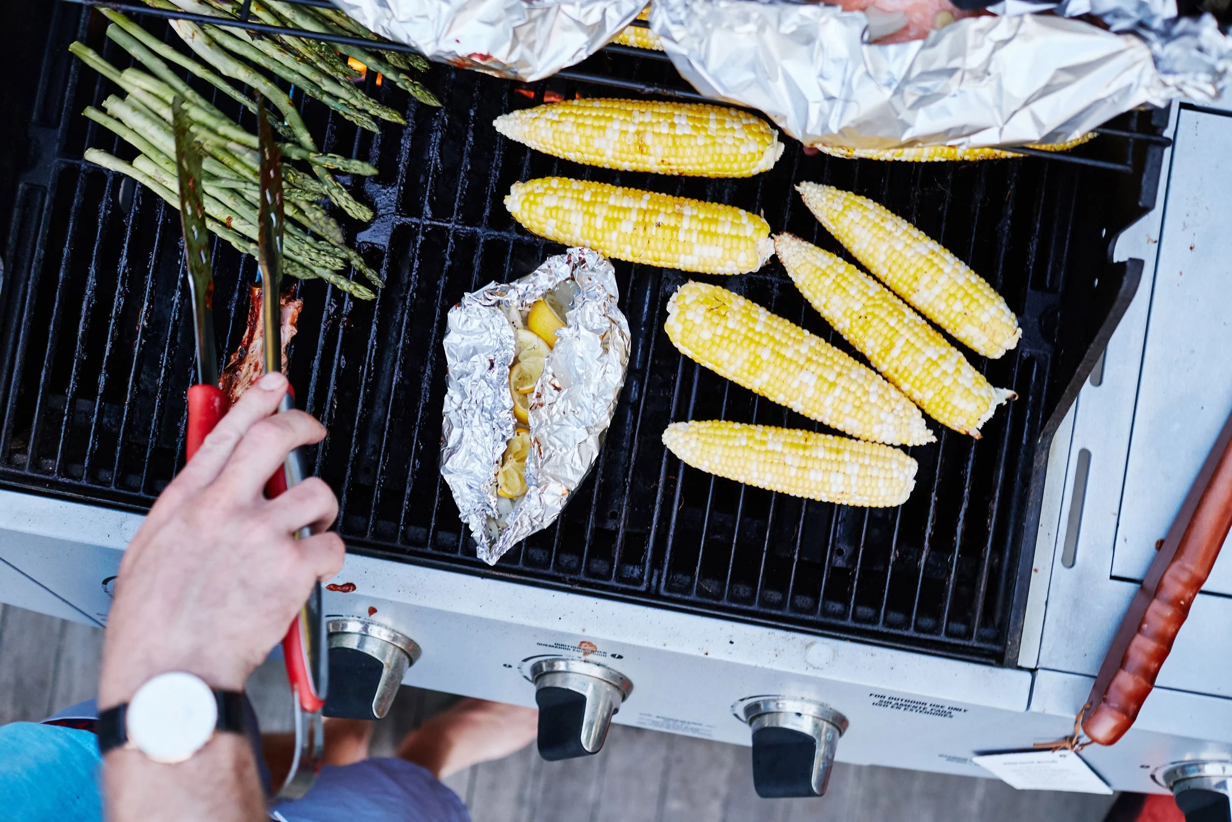 Person grilling corn, asparagus, and foil-wrapped food on a barbecue.