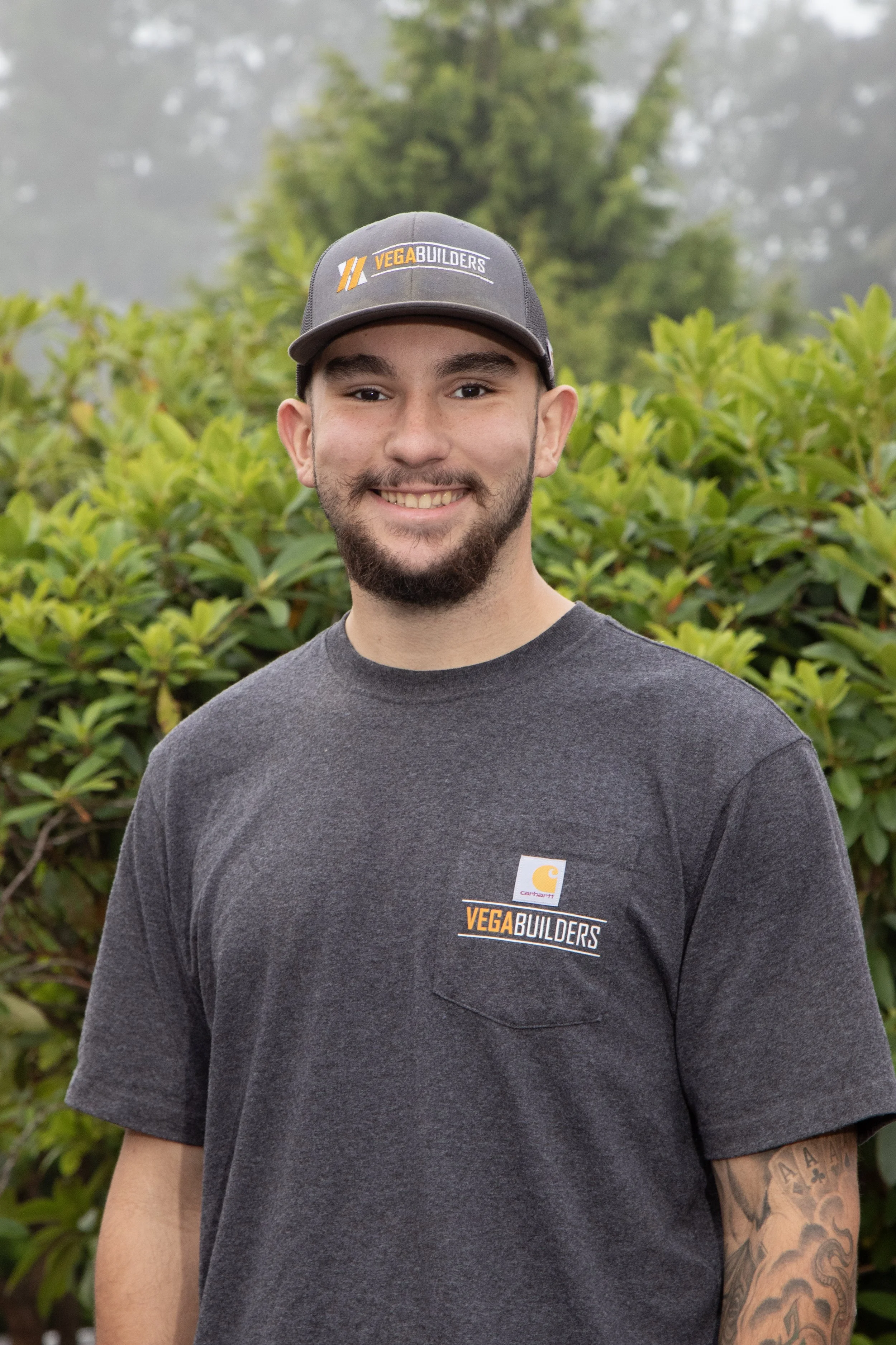 A smiling man with a beard wearing a dark baseball cap with the 'VEGA BUILDERS' logo and a dark hoodie with matching logo, standing outdoors with autumn-colored trees and bushes in the background.