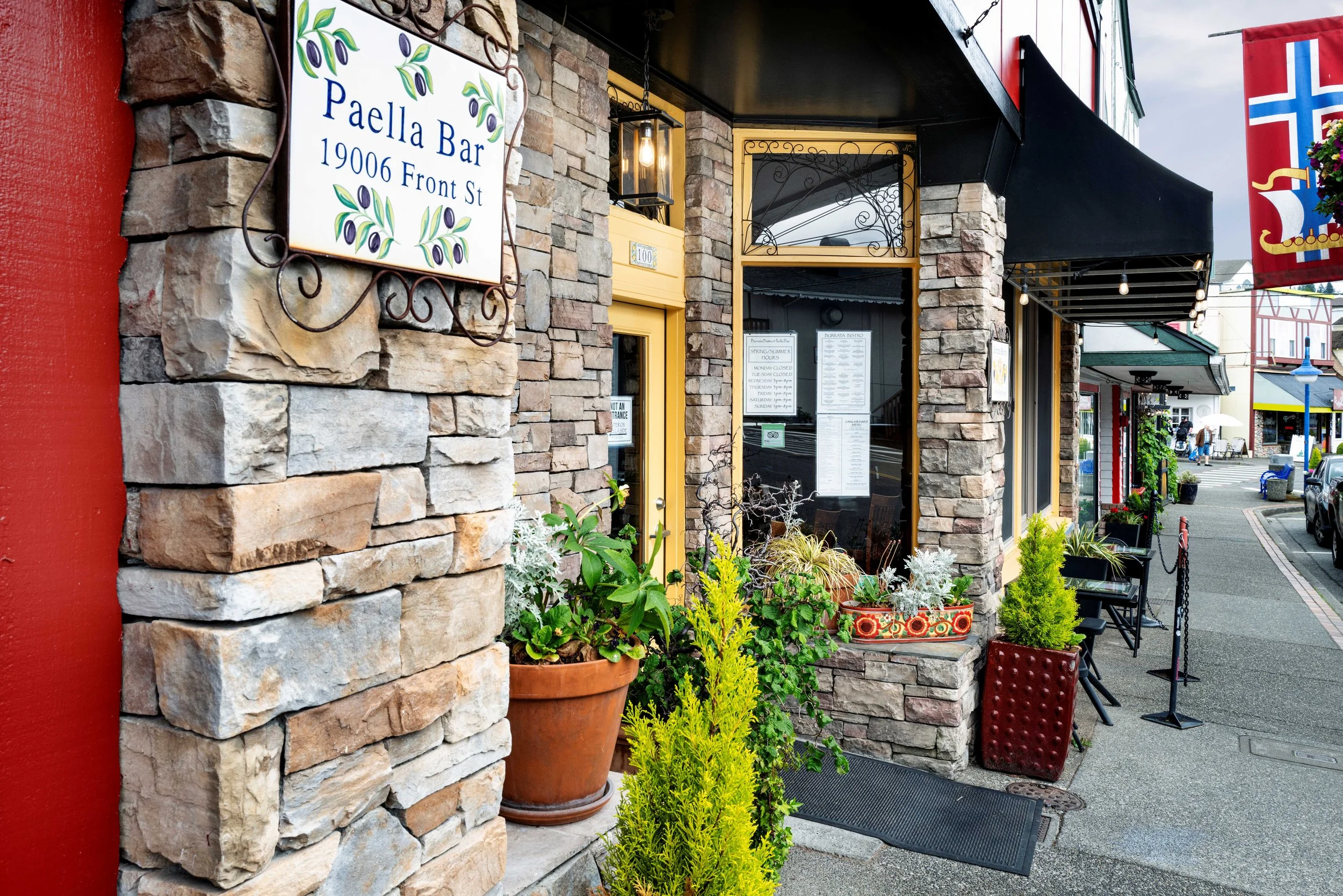 Exterior view of a restaurant called Paella Bar with a stone facade, potted plants, and outdoor seating, located at 19006 Front Street. Vega Builders commercial construction, Poulsbo, WA.