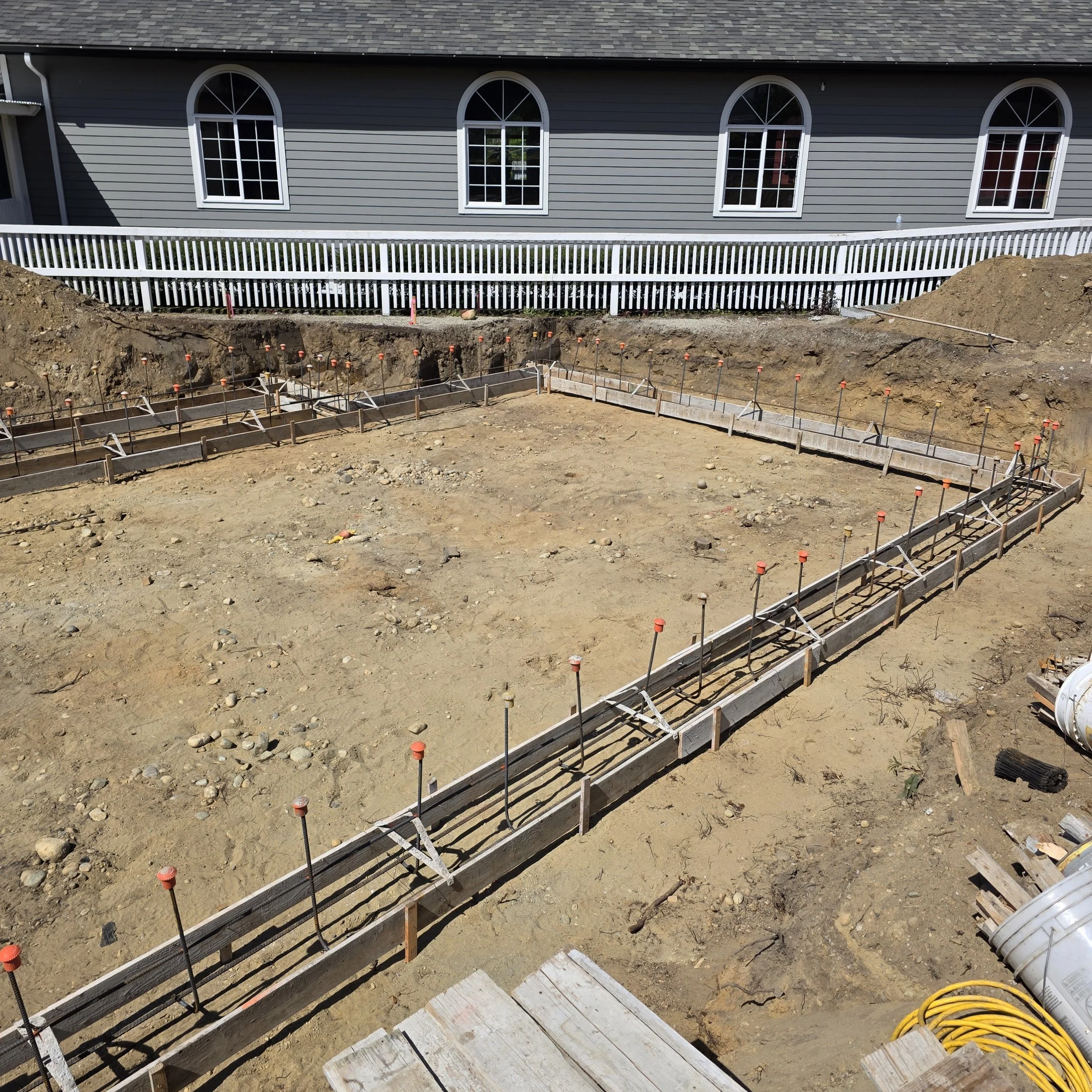Construction site with exposed ground, wooden forms, and orange markers for concrete pouring. In the background is a house with gray siding and white-trimmed windows.