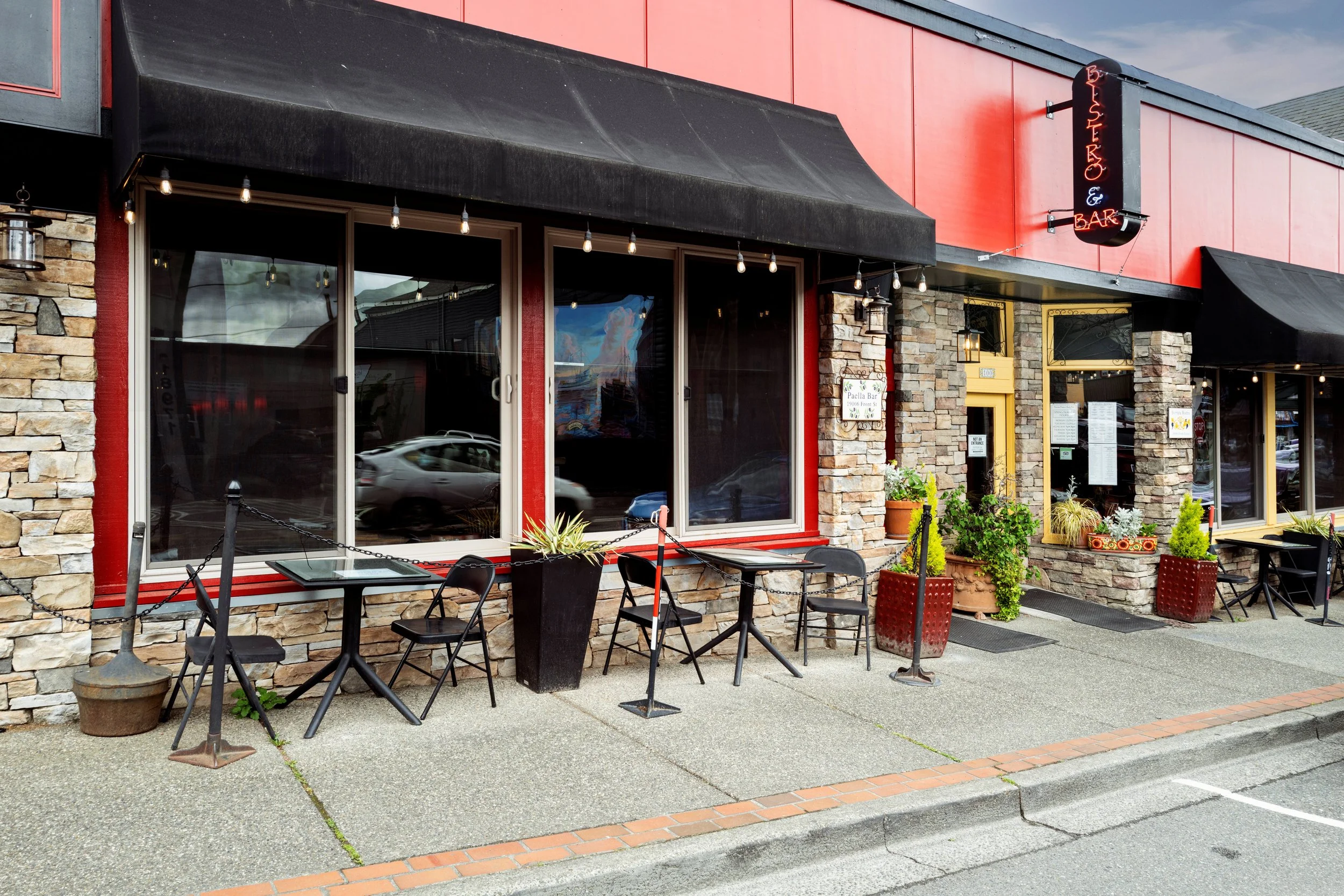 Exterior view of a restaurant with stone and red exterior walls, black awnings, and large windows. There are outdoor tables with chairs and potted plants on the sidewalk. A neon sign on the building reads "Beniso E Bar."