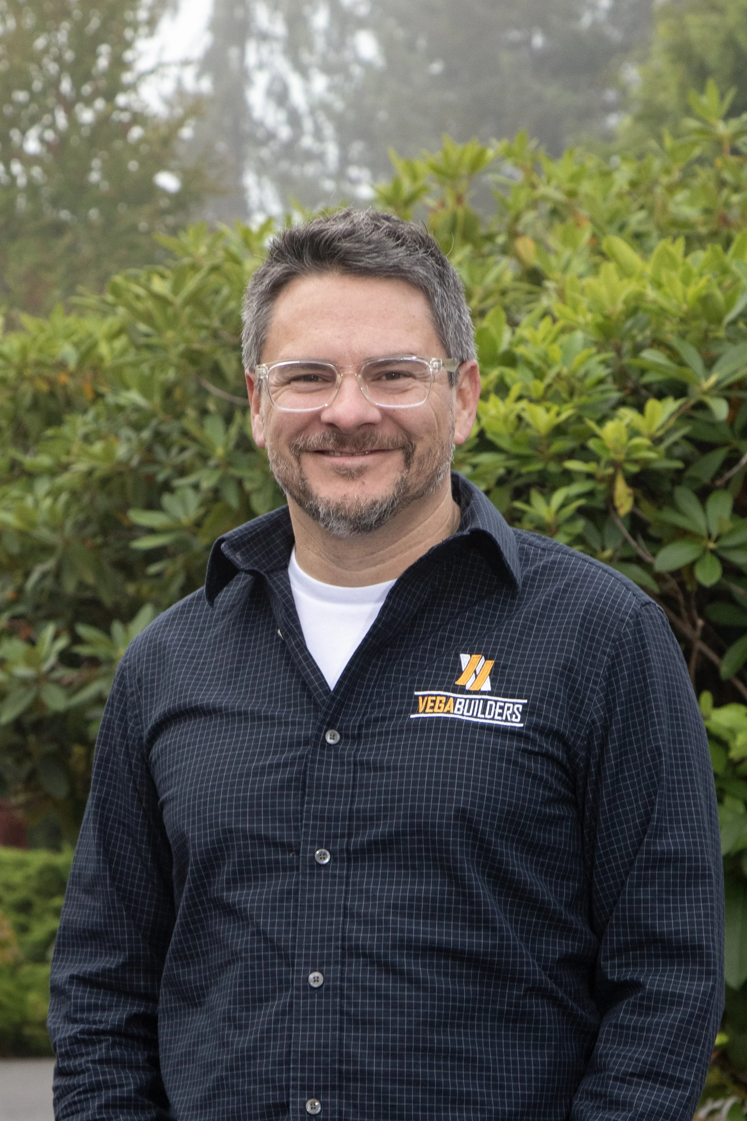 A smiling man with glasses and short dark hair, wearing a black jacket with a 'Vega Builders' logo, outdoors with blurred autumn foliage in the background.
