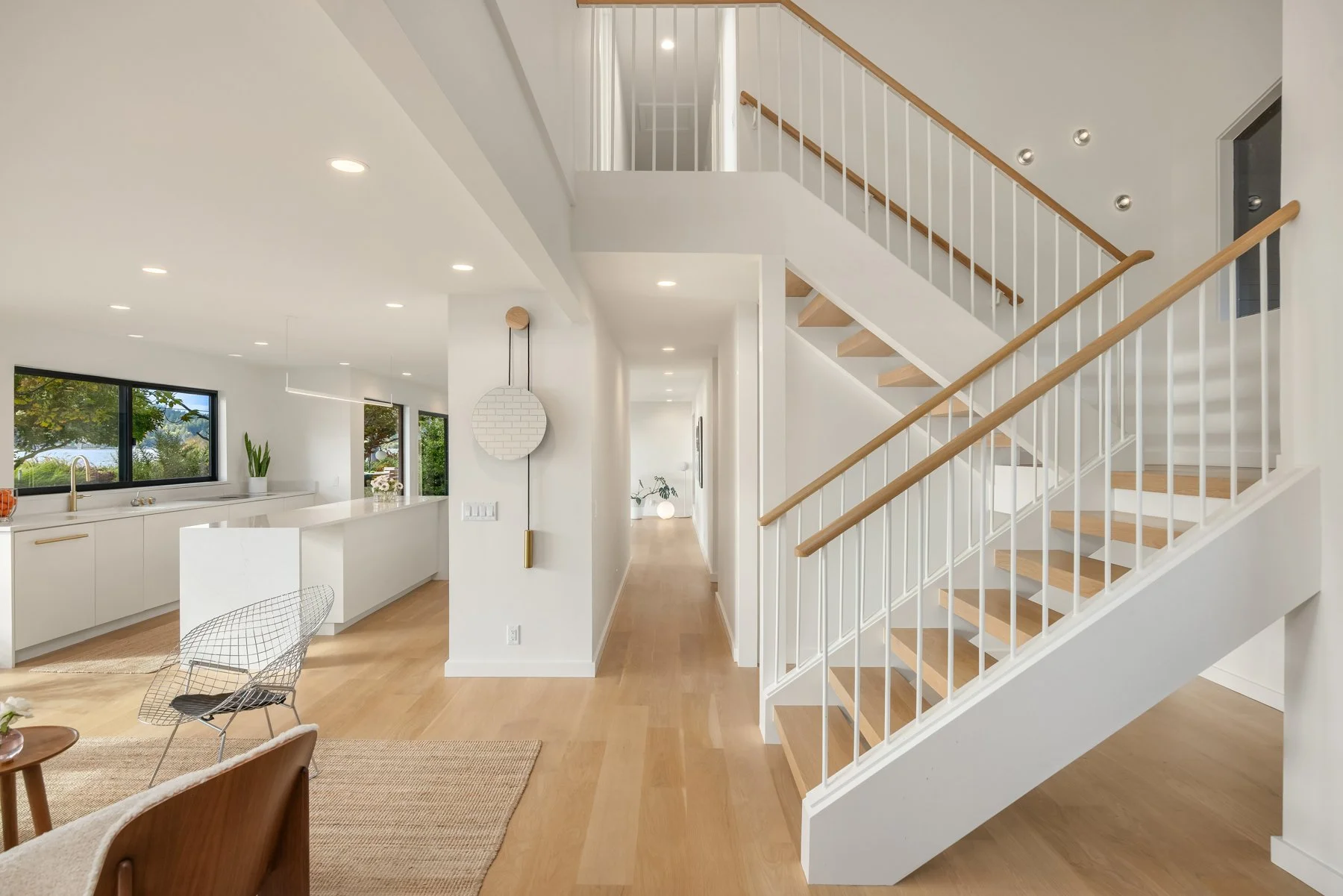 Modern open-concept kitchen and living space with wooden flooring, white walls, staircase with white railings and wooden handrails, large window showing greenery outside, and minimal decor. Vega Builders home remodeling and construction.