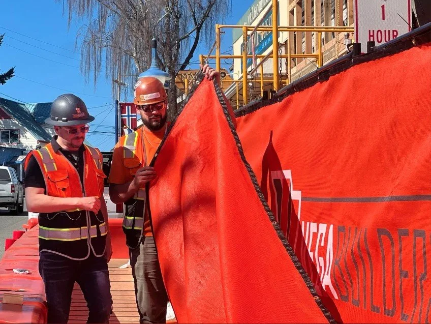 Two construction workers, wearing safety helmets and vests, hold an orange safety barrier on a street.