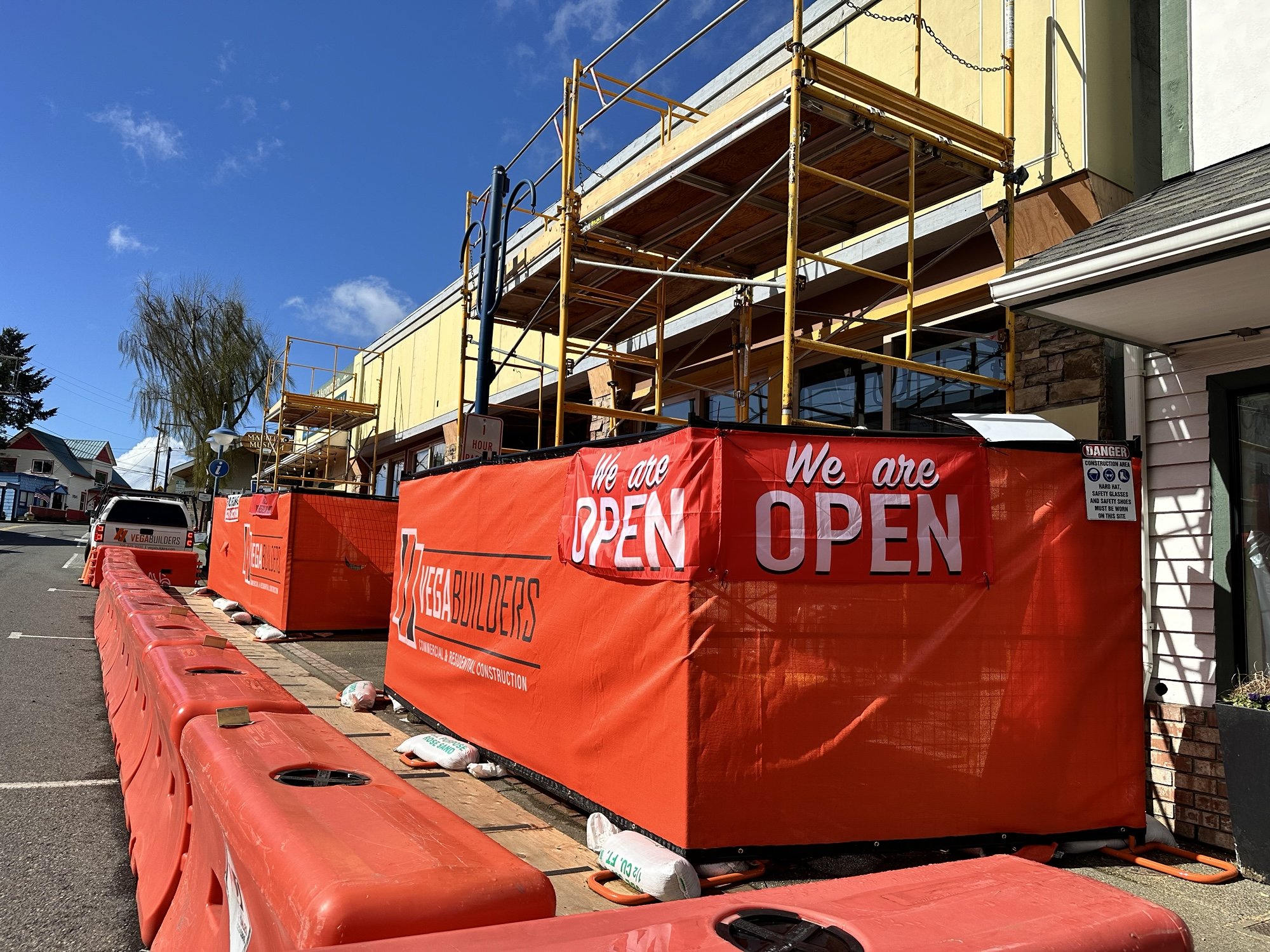 Construction site with scaffolding and red barriers in front of a building with yellow and green walls. There is a sign that says 'We are OPEN' and another sign indicating safety rules.