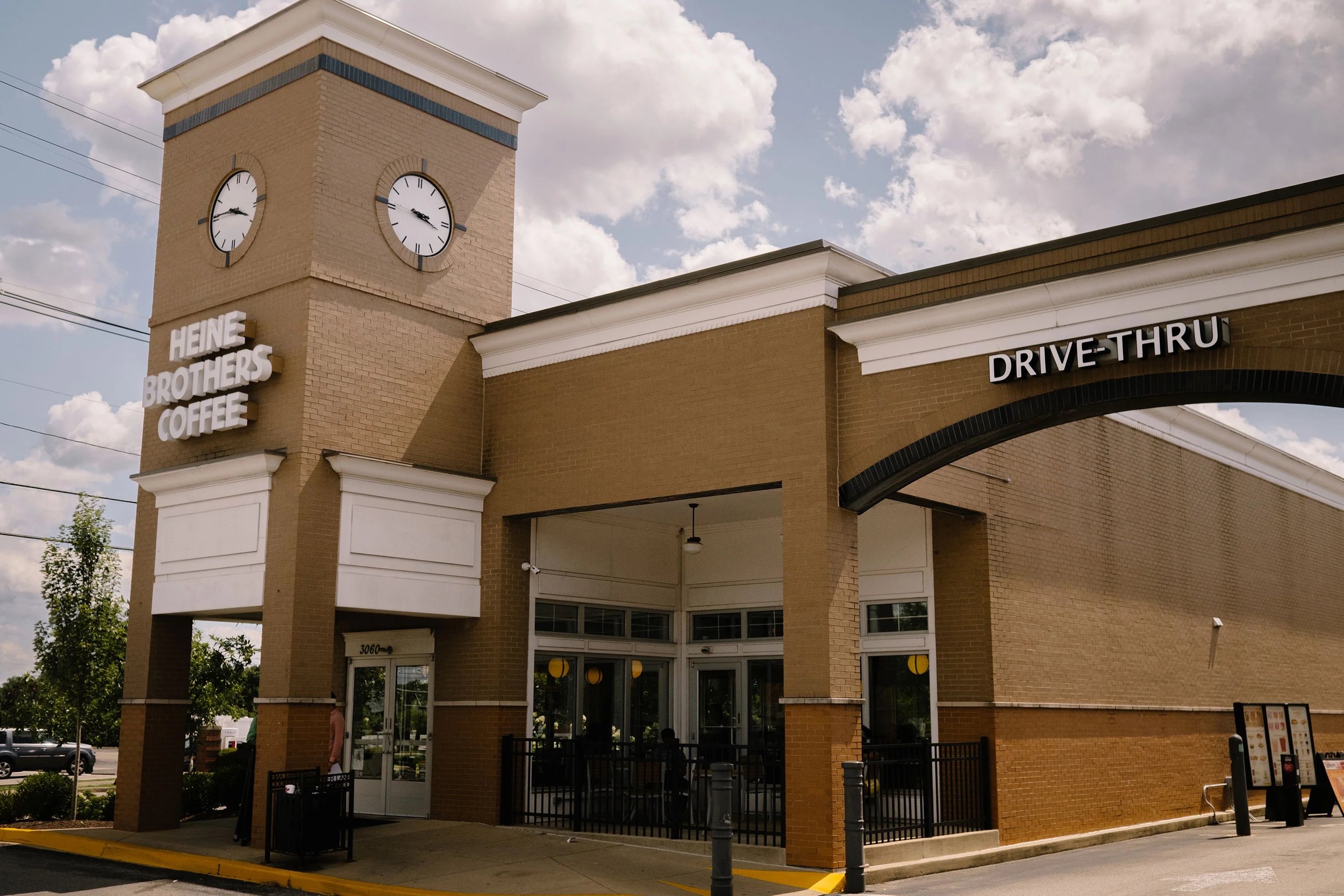 Exterior of a café or restaurant with large windows, black and brown wooden facade, small trees in planters, and a neon sign inside that reads 'Heine Brothers'.