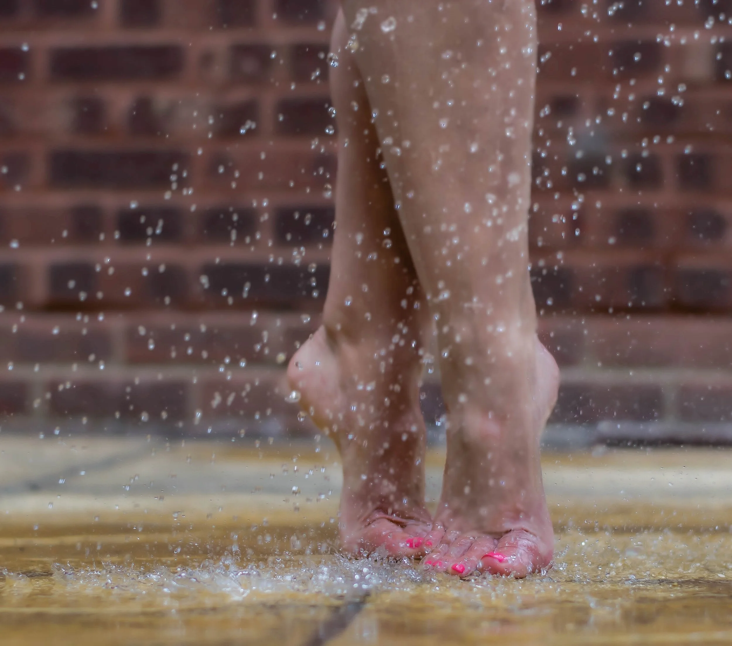 Close-up of bare feet walking on wet ground with water splashing, with a brick wall in the background.