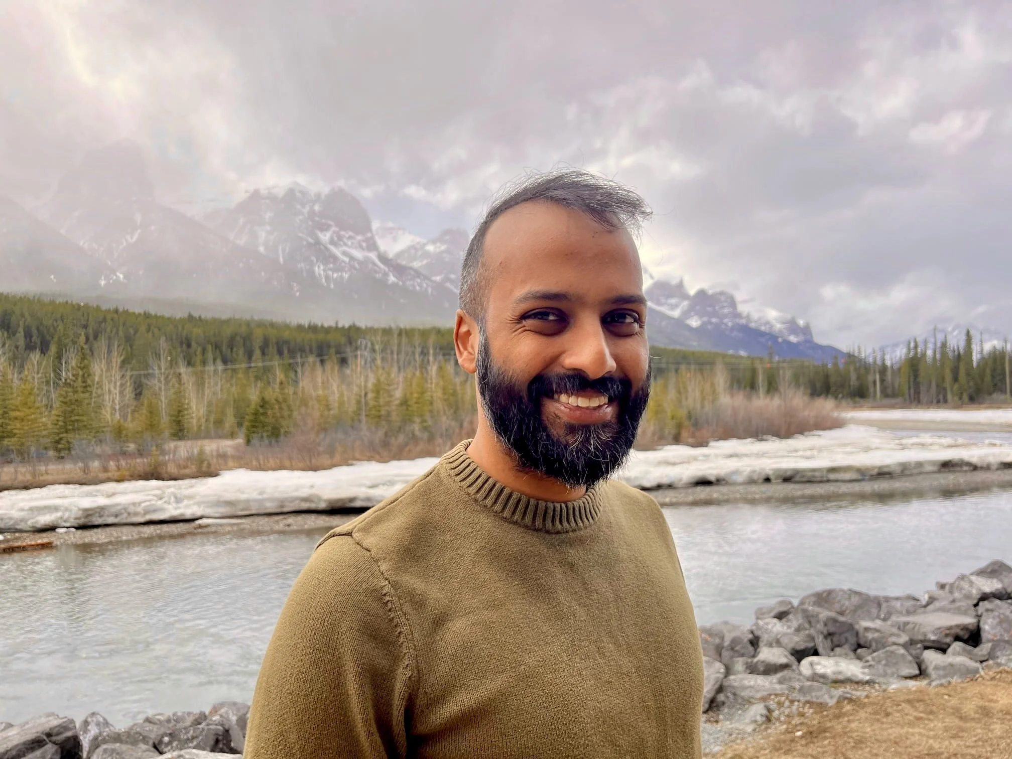 A smiling man who is a psychologist in Alberta, Canada, with a beard and short hair stands outdoors by a river, with mountains, evergreen trees, and a cloudy sky in the background.