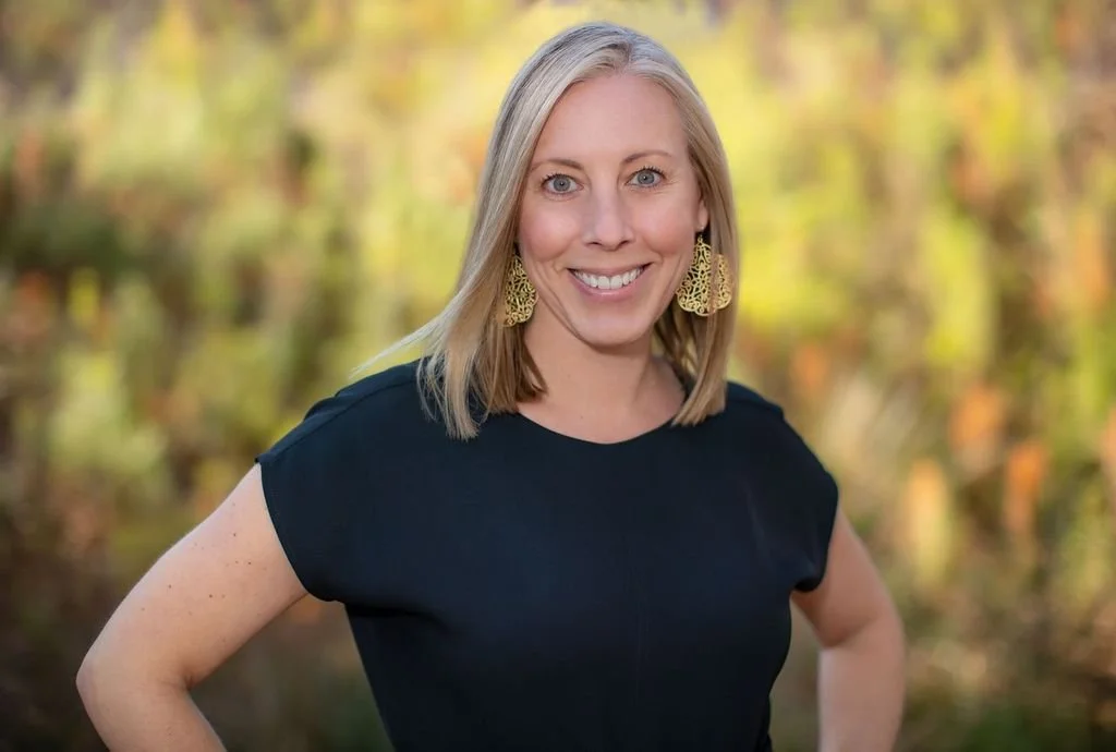 A woman with blonde hair and blue eyes smiling outdoors with a blurred autumnal background, wearing a black top and large gold earrings.