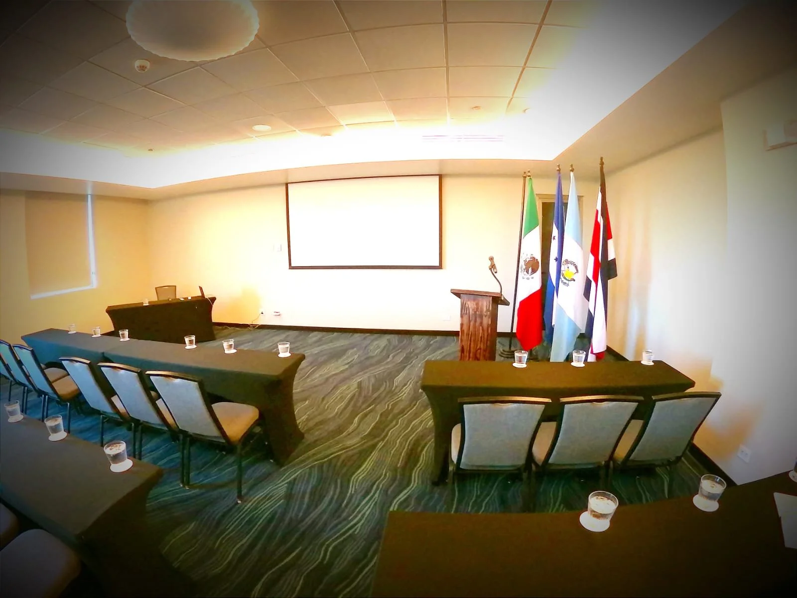 Sala de conferencia con una pantalla grande, bandera de México, y otros países, varias mesas con sillas y velas encendidas, preparado para un evento formal.