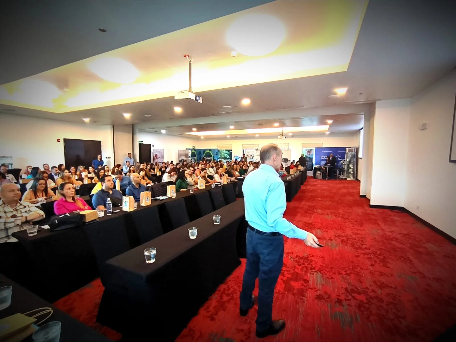 Conferencia en un salón con muchas personas sentadas en mesas y un presentador de pie frente a la audiencia.