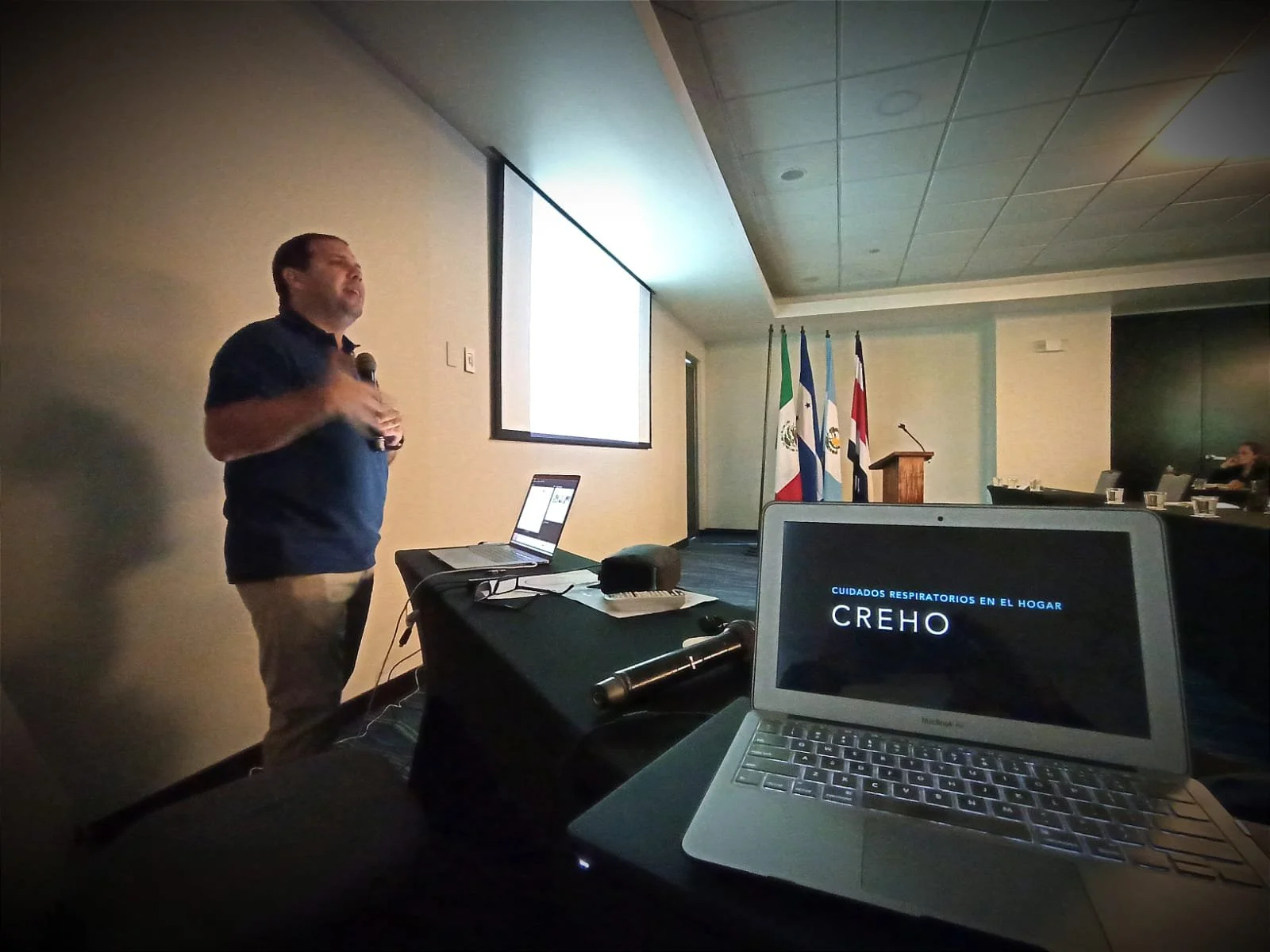 Hombre dando una plática o conferencia en un salón, sosteniendo un micrófono, con una pantalla grande detrás y banderas en el fondo. Una laptop en la mesa muestra la palabra 'CREHO' en la pantalla.