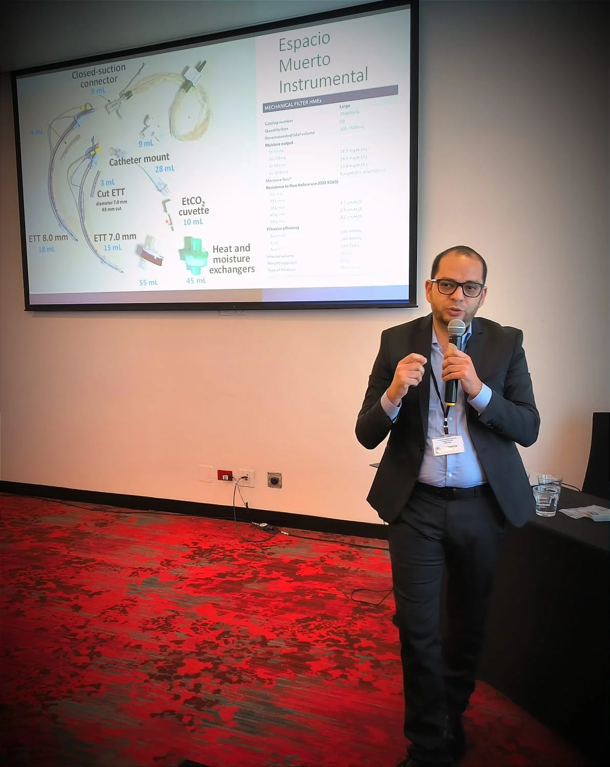 Hombre de negocios dando una presentación en una conferencia, con una diapositiva de equipo médico en una pantalla detrás.